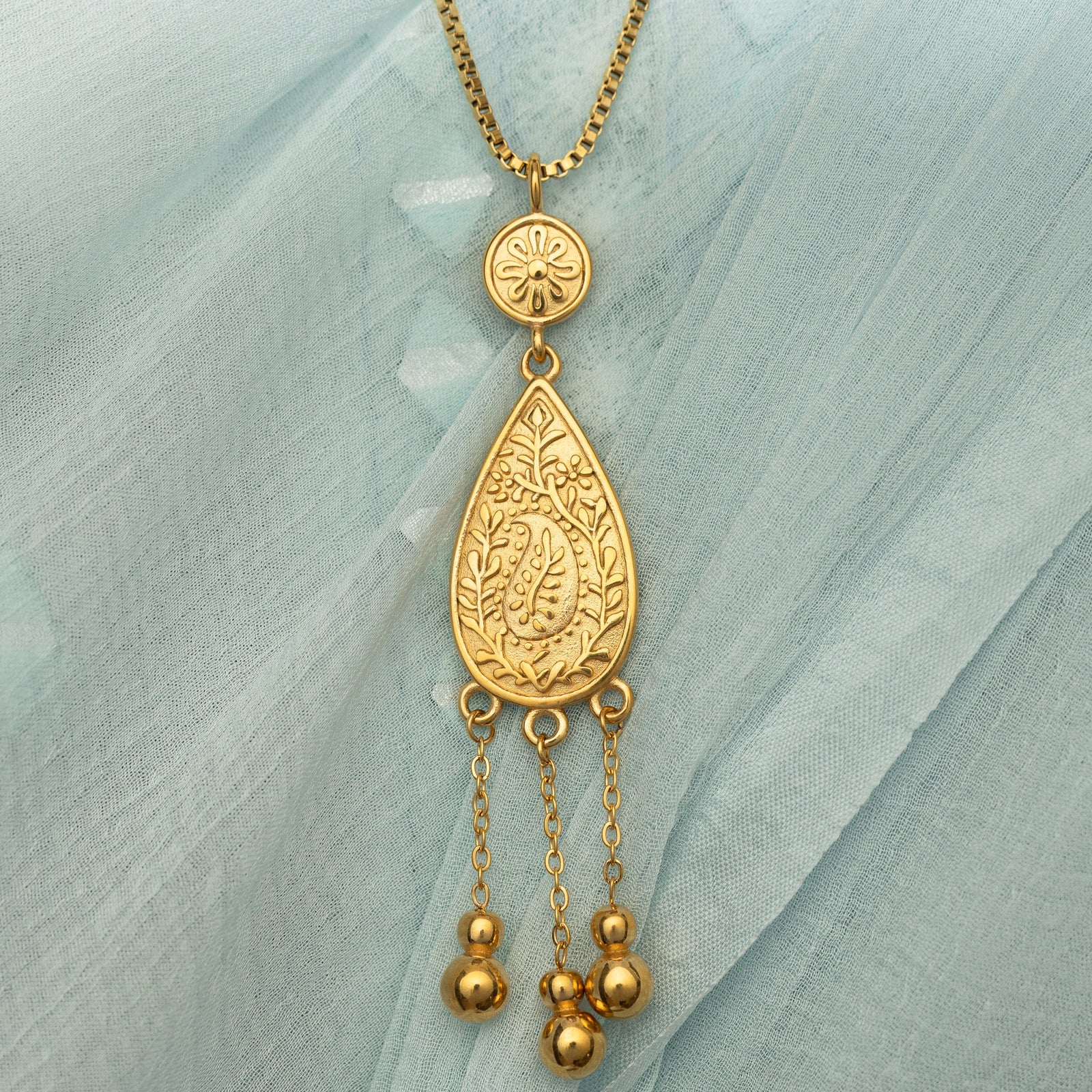 Close up photo of the gold pendant on a light blue fabric background.