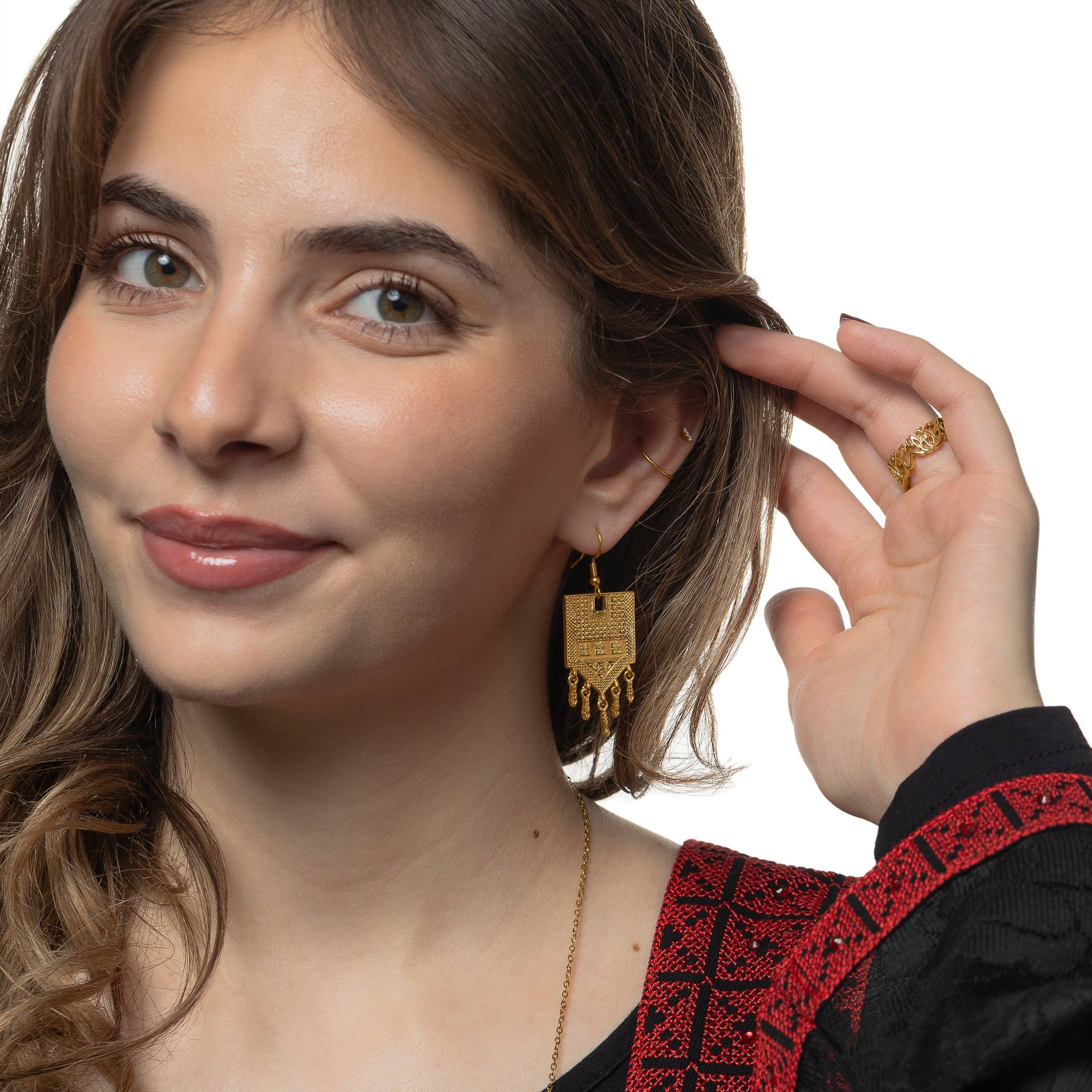 Photo of the model smiling to the camera, holding their hair away from their left ear. The model is wearing the gold Raining Tatreez Earrings