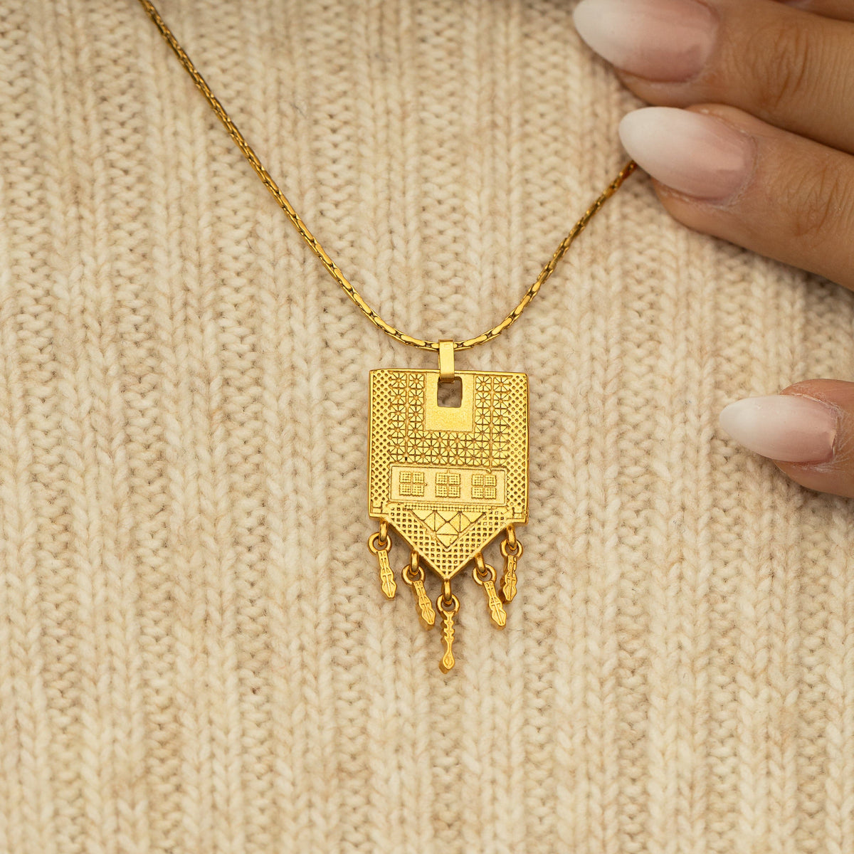 Close-up of the Raining Tatreez Necklace resting on a cream knit sweater, showing the detailed patterns and dangling charms on the gold pendant.
