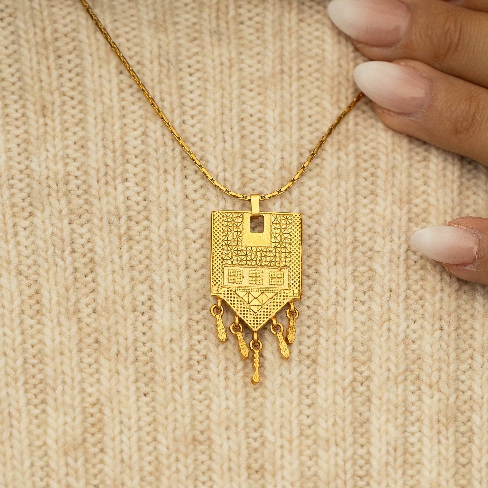 Close-up of the Raining Tatreez Necklace resting on a cream knit sweater, showing the detailed patterns and dangling charms on the gold pendant.