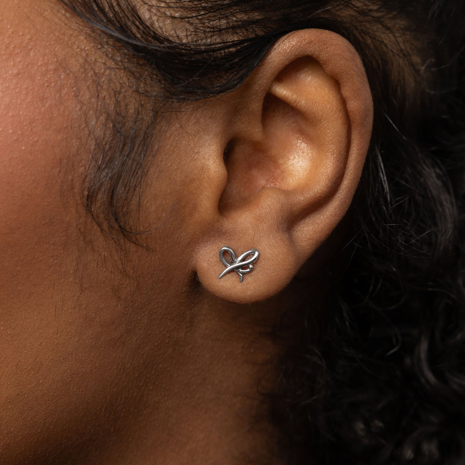 A female model with medium skin tone showcasing the silver "Mama" Ribbon Studs. The earrings rest elegantly on her earlobes, enhancing her simple attire and drawing attention to the detailed ribbon motif.