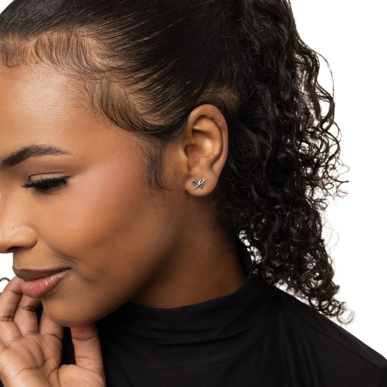 A female model with medium skin tone showcasing the silver "Mama" Ribbon Studs. The earrings rest elegantly on her earlobes, enhancing her simple attire and drawing attention to the detailed ribbon motif.