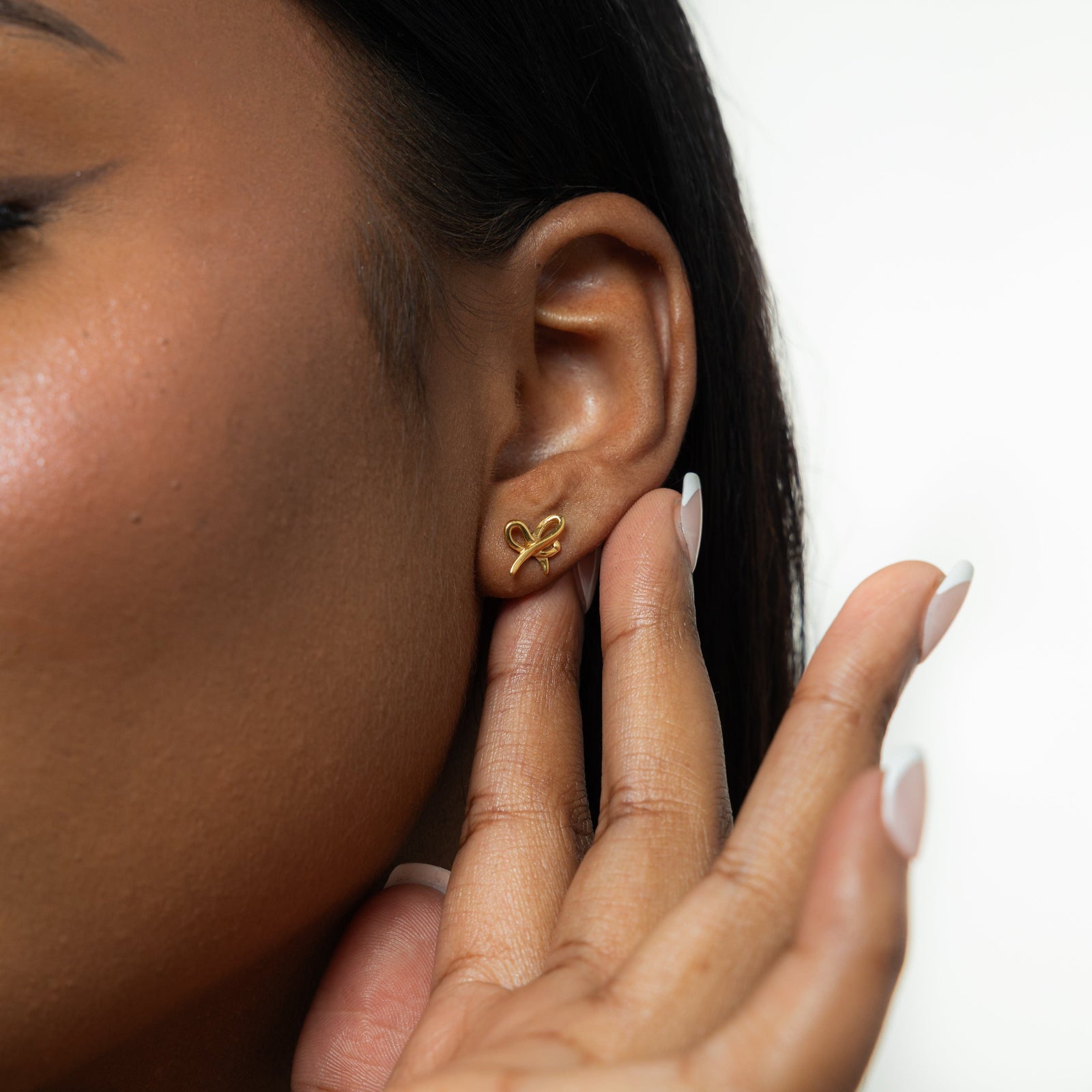 A female model with medium skin tone wearing the 18K gold-plated "Mama" Ribbon Studs. The earrings sit delicately on her earlobes, complementing her minimalist outfit and highlighting the intricate ribbon design.