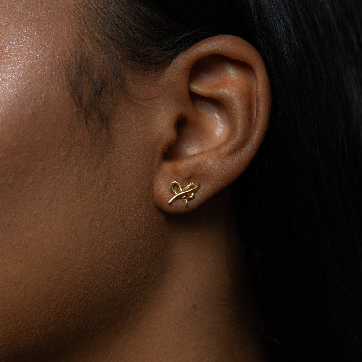 A female model with medium skin tone wearing the 18K gold-plated "Mama" Ribbon Studs. The earrings sit delicately on her earlobes, complementing her minimalist outfit and highlighting the intricate ribbon design.