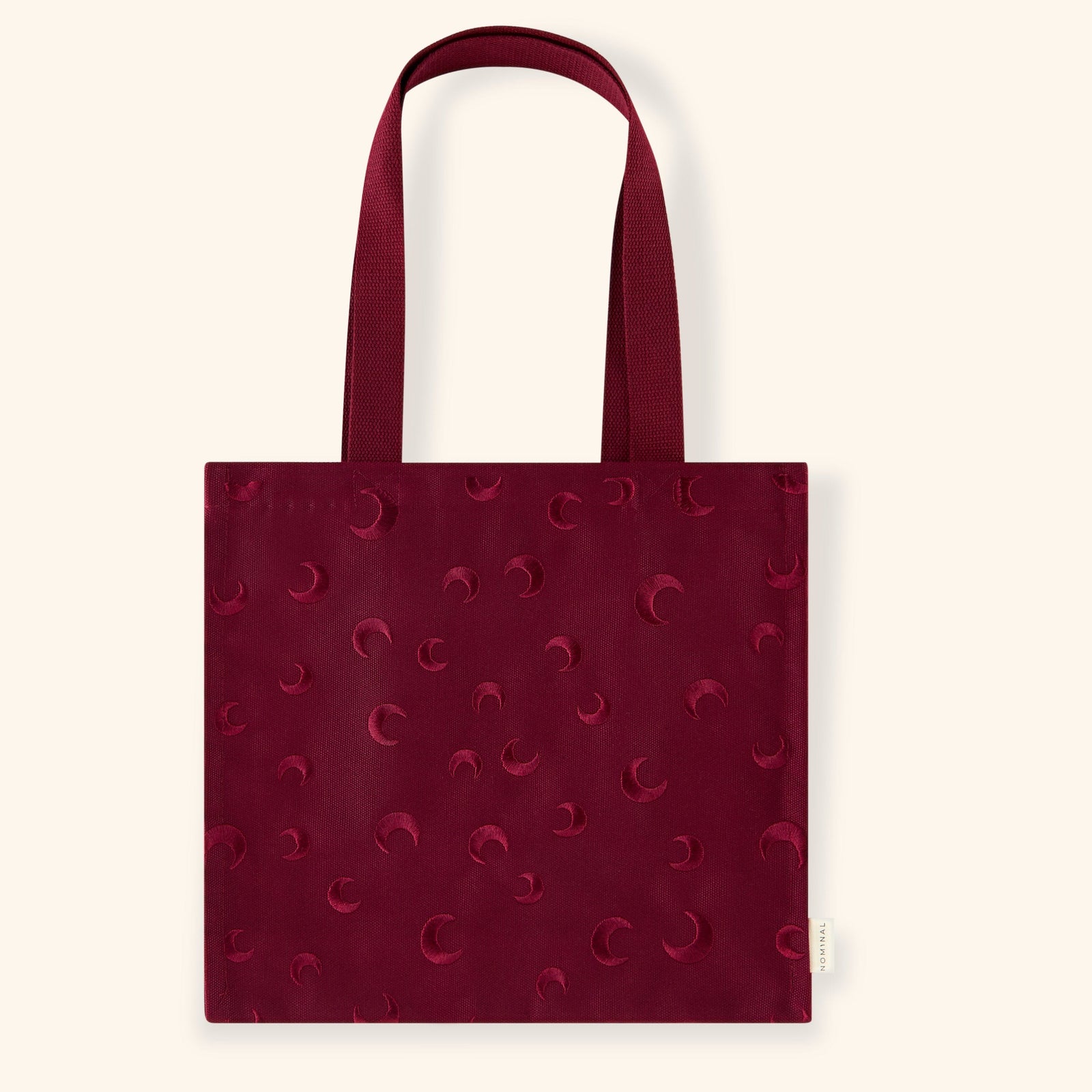 Deep maroon skyfall tote bag with an all-over embroidered pattern of crescent moons in matching maroon thread. Made from sturdy fabric with a subtle textured finish.