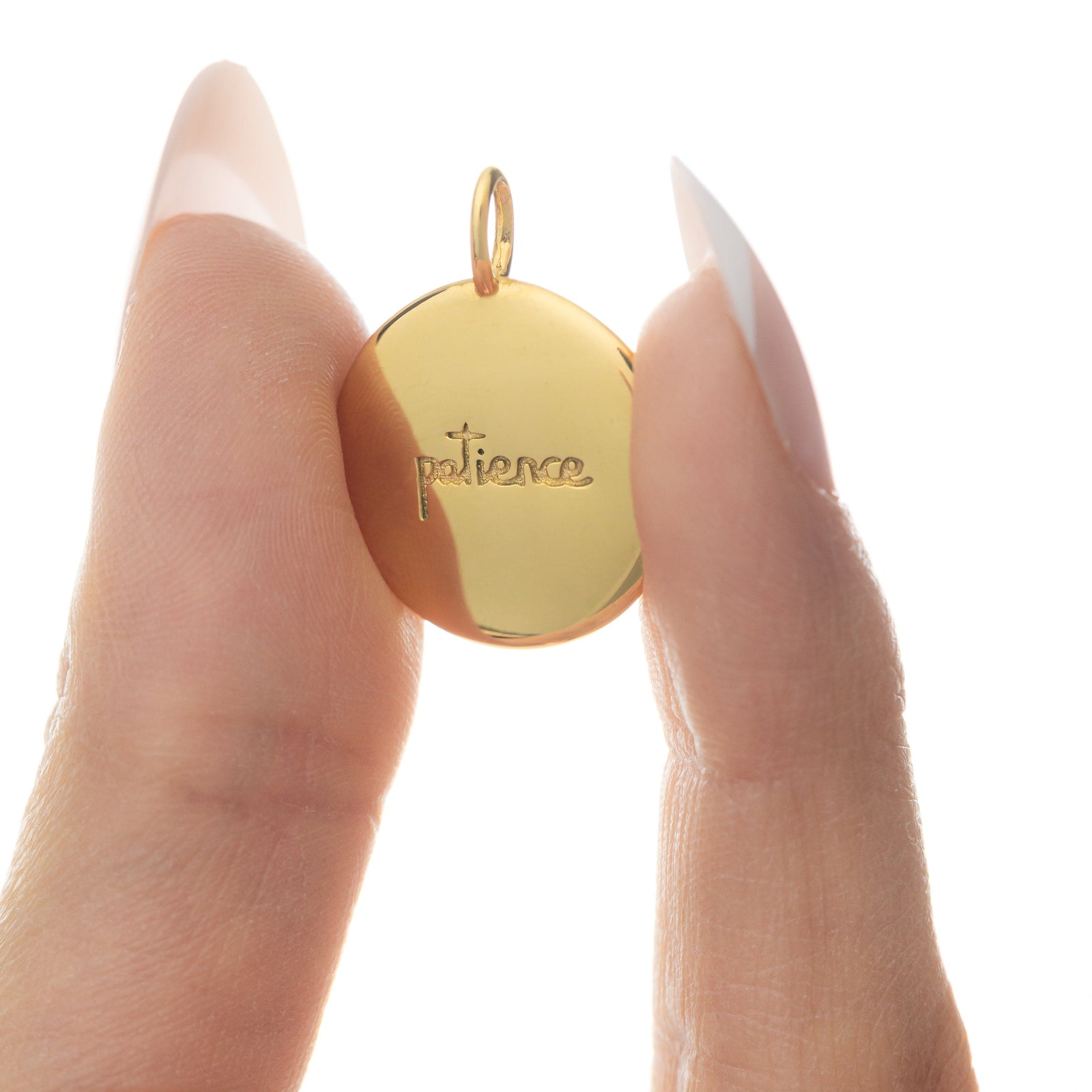 Fingers holding the gold Sabr charm from the reverse side, displaying the English word "patience" engraved in cursive.