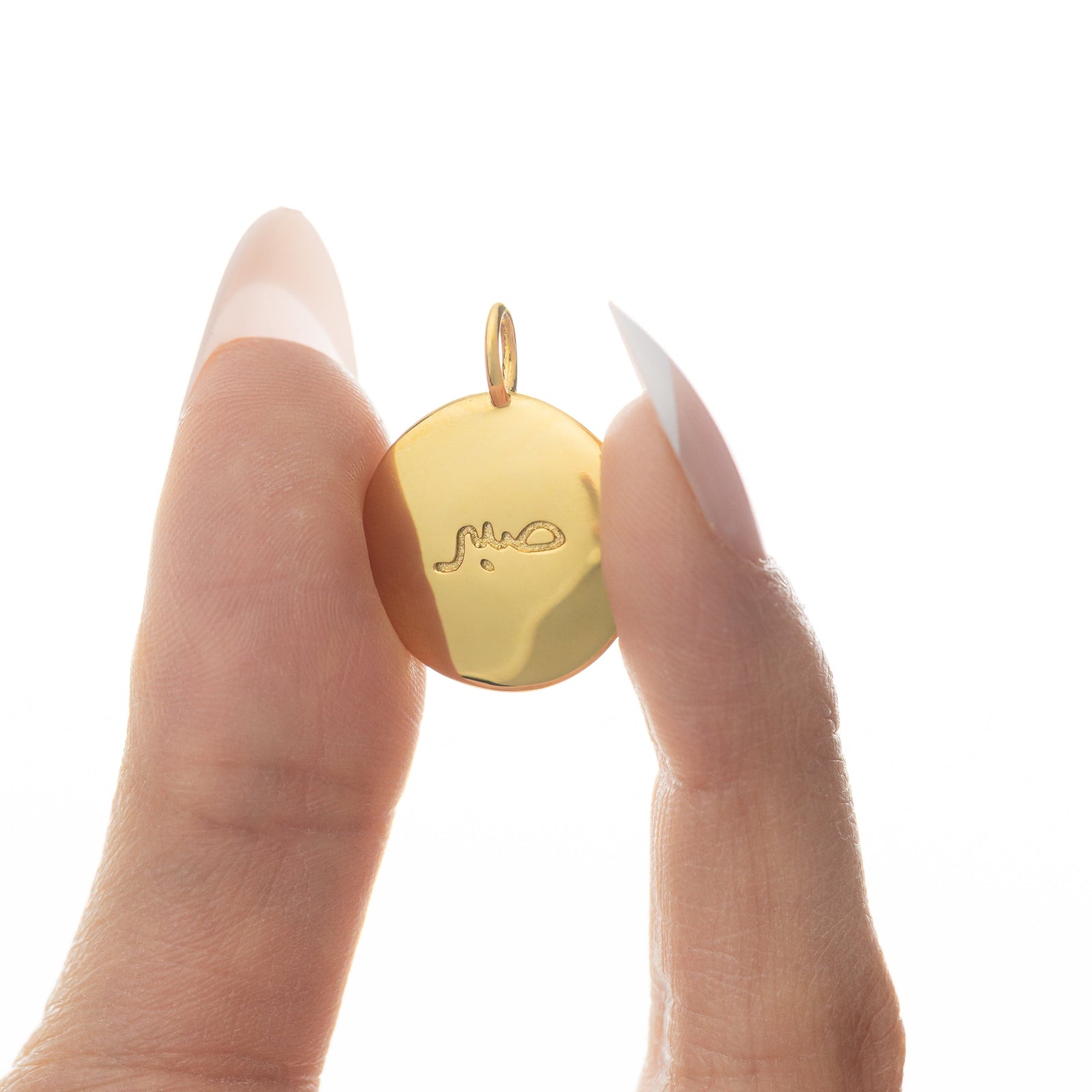 Fingers holding the gold Sabr charm up close, showing the Arabic engraving.