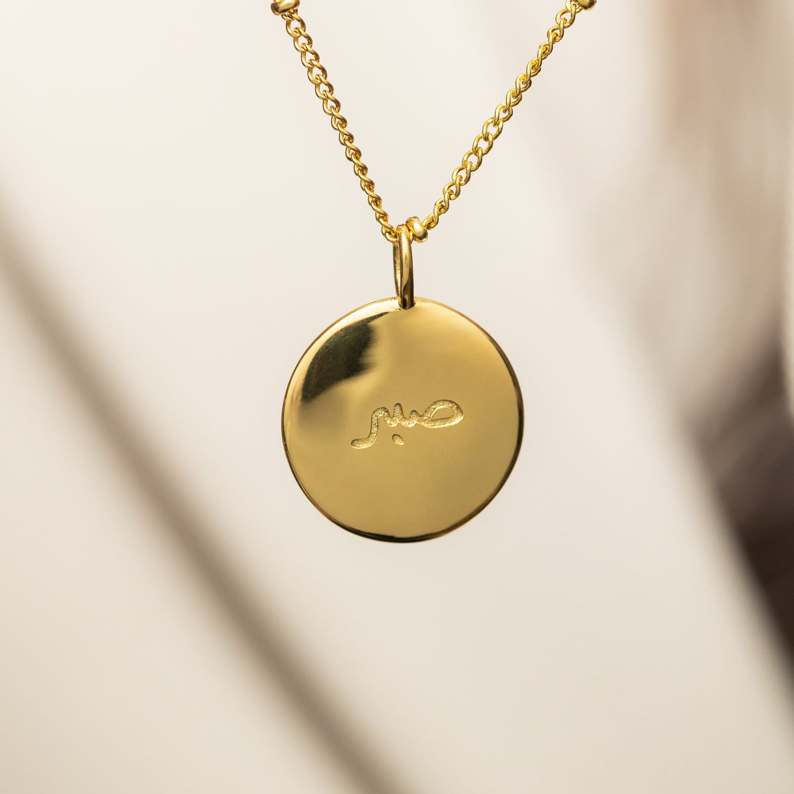 Gold necklace with round "Sabr" charm hanging from a polished chain, shot against a softly blurred neutral background.