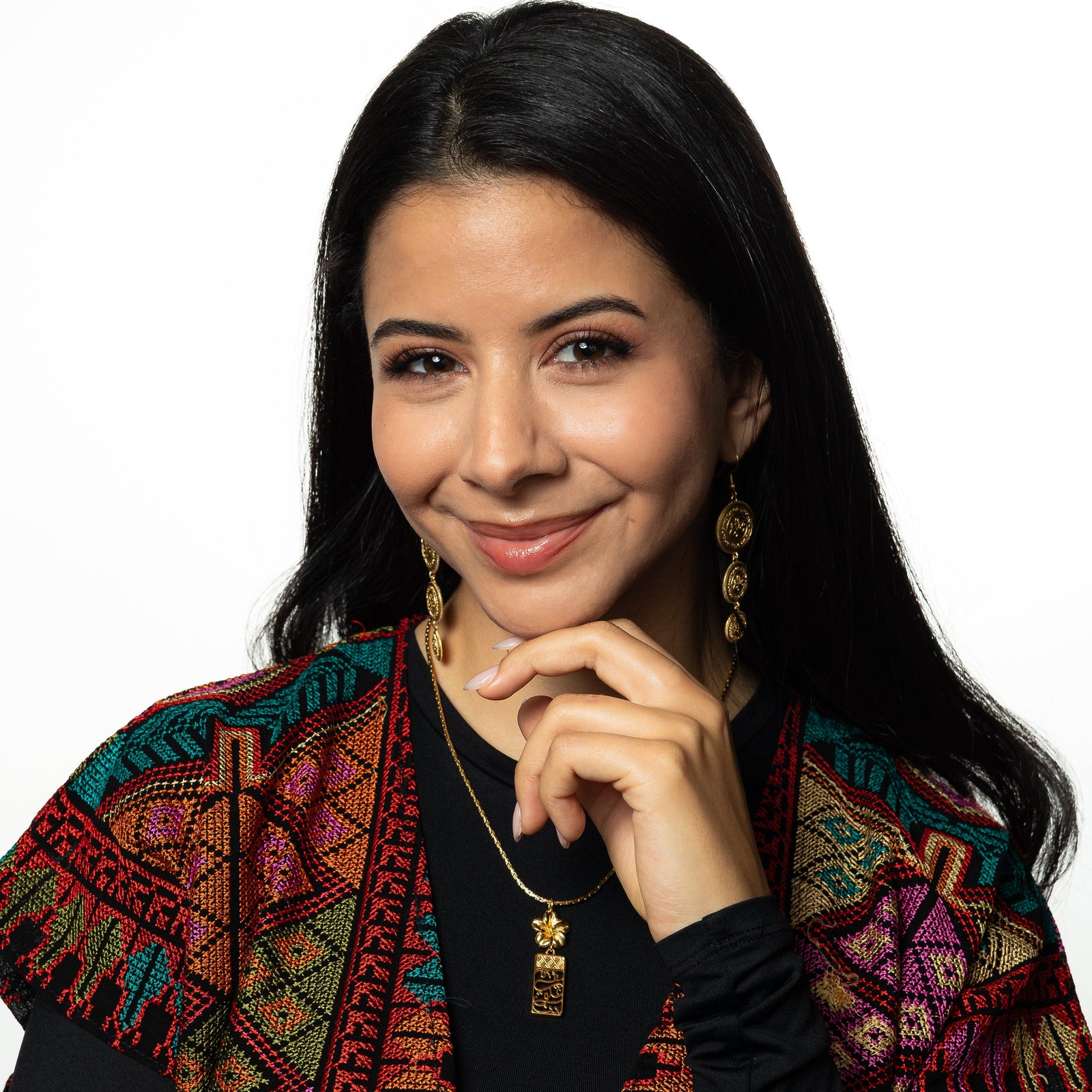 Photo of the model smiling at the camera, touching their chin with their left hand. The model is wearing a pair of gold Sabr | Patience Coin and gold Blooming In Our Hearts Necklace.