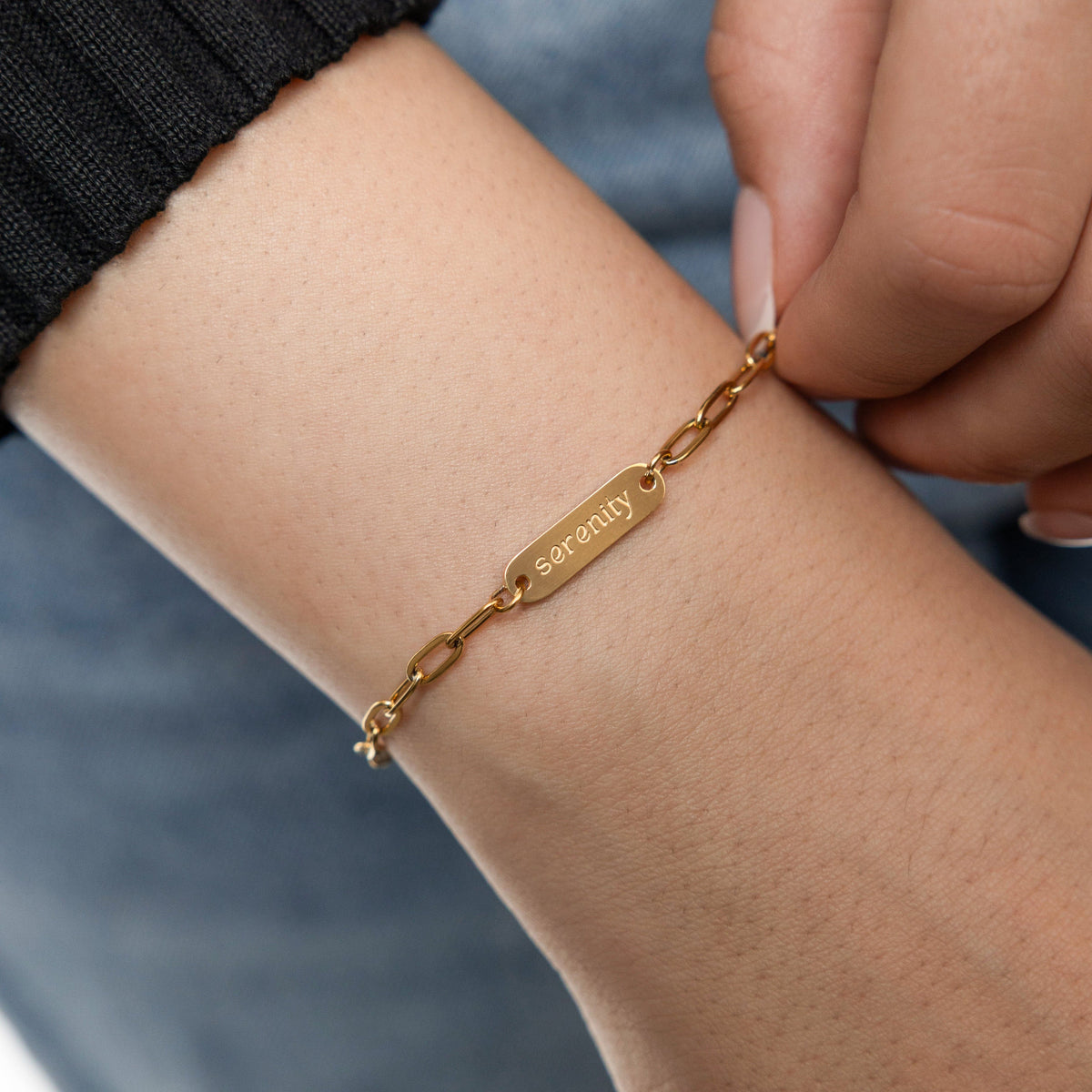 The image is a close-up photograph of a person’s wrist wearing a delicate gold bracelet. The bracelet features a thin chain with a small rectangular plate in the center. Engraved on the plate is the word “Serenity,” which stands out subtly against the shiny gold surface. The bracelet appears minimalistic and elegant, designed as a personalized piece of jewelry.