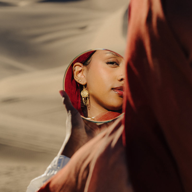 Woman’s reflection in a handheld mirror showing her wearing gold Salam Vintage Token Earrings pimage_ set against a sandy desert background.