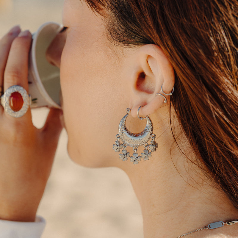 Silver Levantine Garden Hoop Earring with floral engraving and dangling charms worn on a woman’s ear as she holds a cup.
