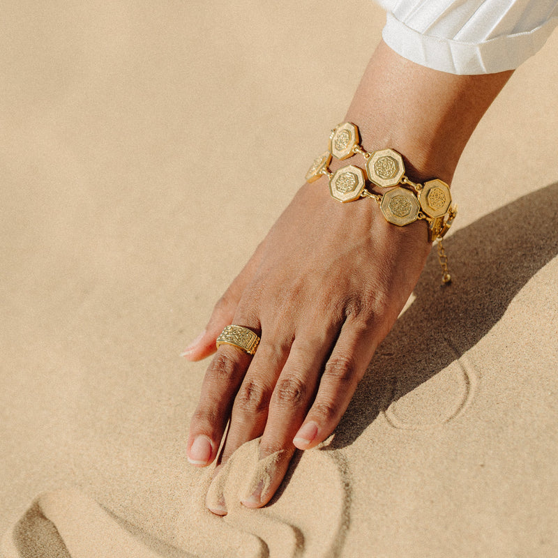 The “Our Roots Run Deep” Dotted Bracelet in gold worn on the wrist resting on sand.