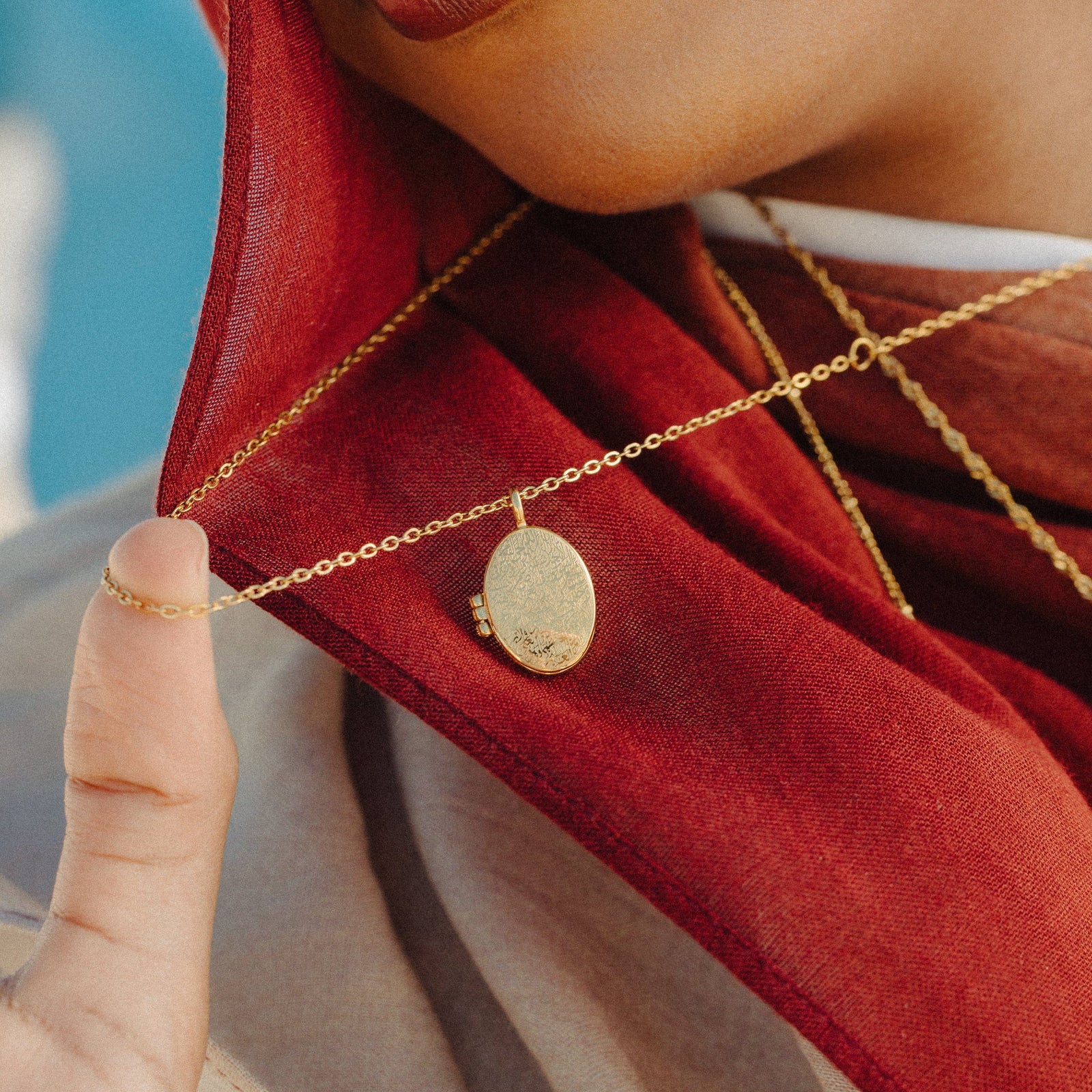 Gold Ayatul Kursi Locket worn over a layered red scarf, with a hand lifting the chain to show the engraved front of the locket.
