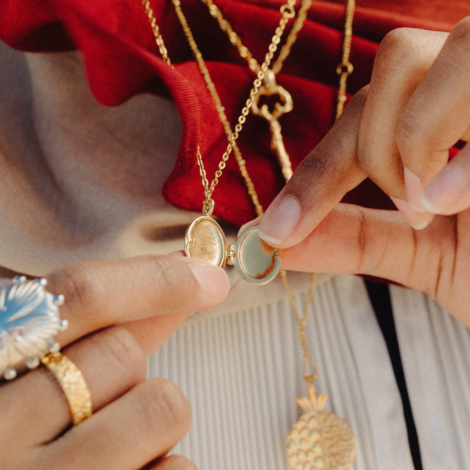 Close-up of hands opening the Ayatul Kursi Locket to show its photo compartment, with layered gold necklaces and fabric textures in the background.