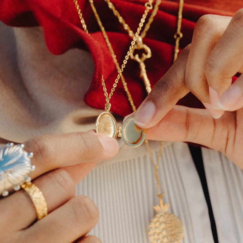 Close-up of hands opening the Ayatul Kursi Locket to show its photo compartment pimage_ with layered gold necklaces and fabric textures in the background.