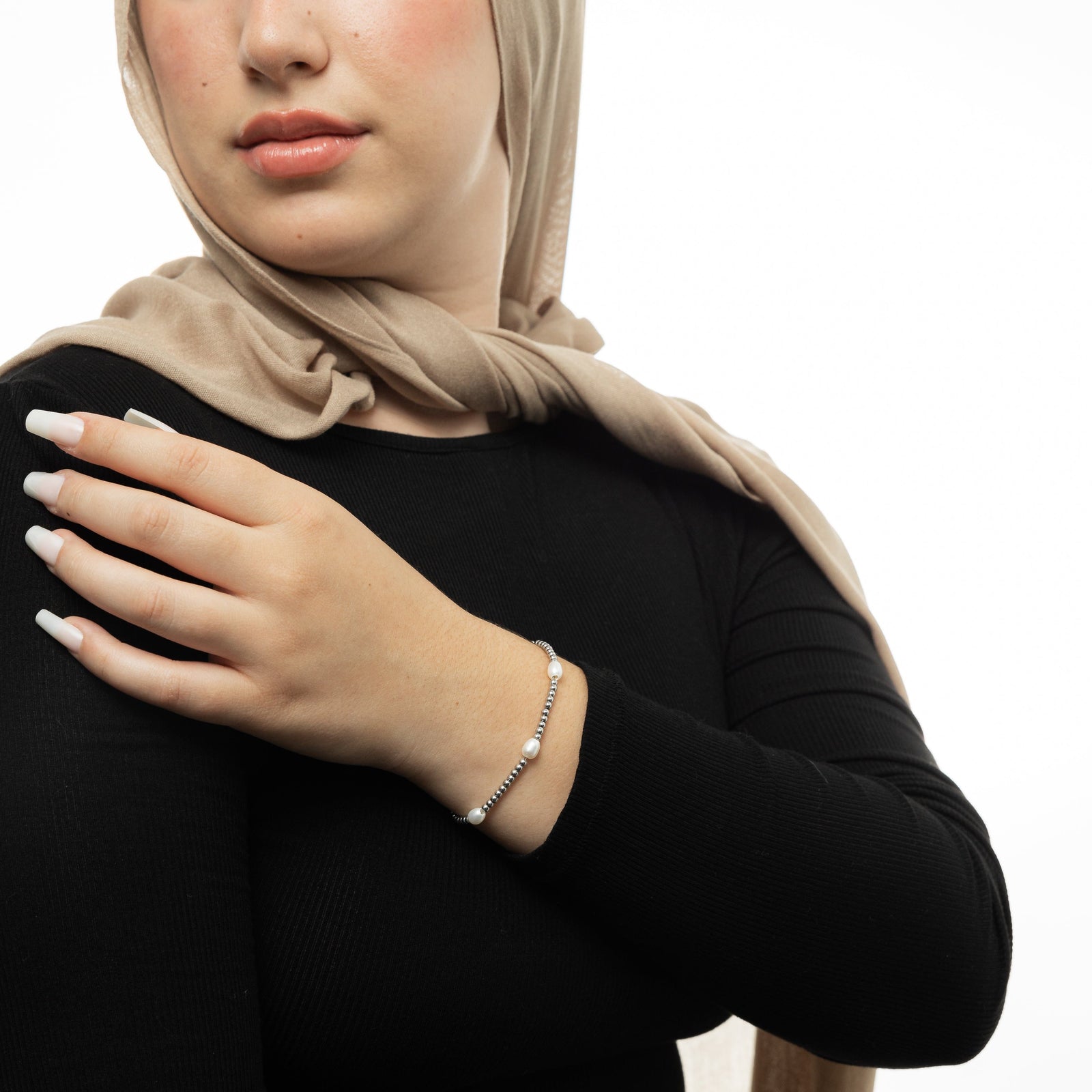 Woman wearing a silver satellite bracelet, styled with a beige hijab, black long-sleeve top, and long white nails, with her hand resting across her chest.