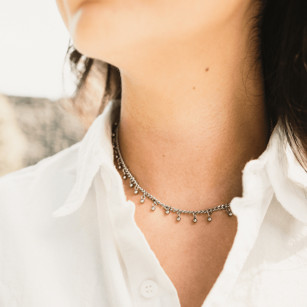 Silver choker with dangling bead accents worn by a woman in a white collared shirt, head turned slightly.