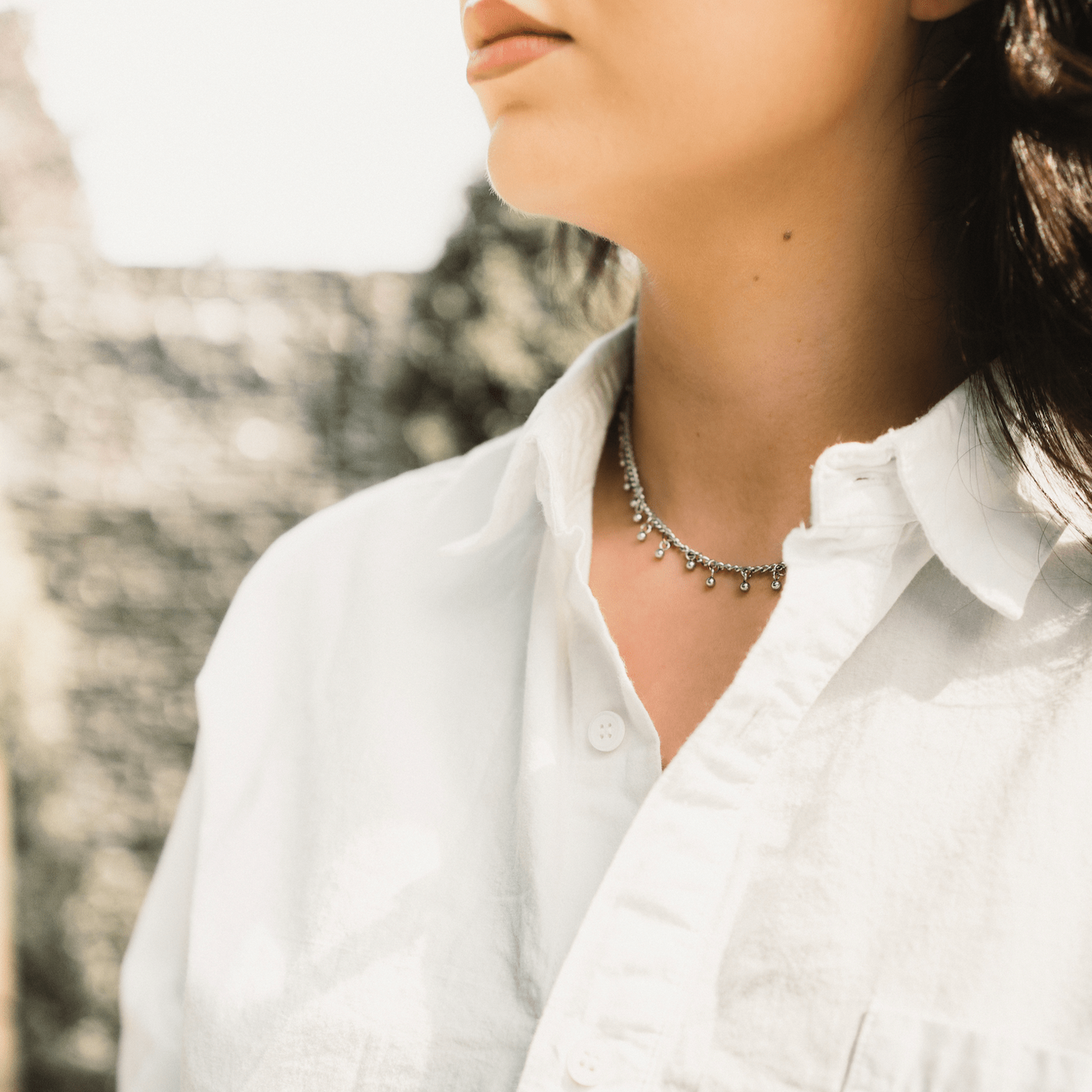 Woman in a white shirt wearing a silver choker with small dangling beads, facing slightly sideways.