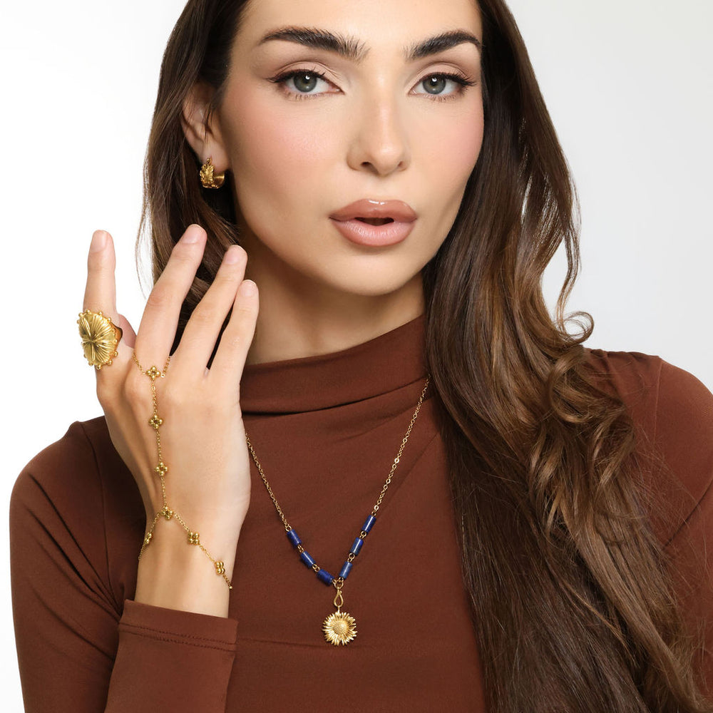 Woman wearing an 18k gold plated necklace with a detailed sunflower pendant and deep blue tube beads, paired with a matching hand chain ring and gold earrings