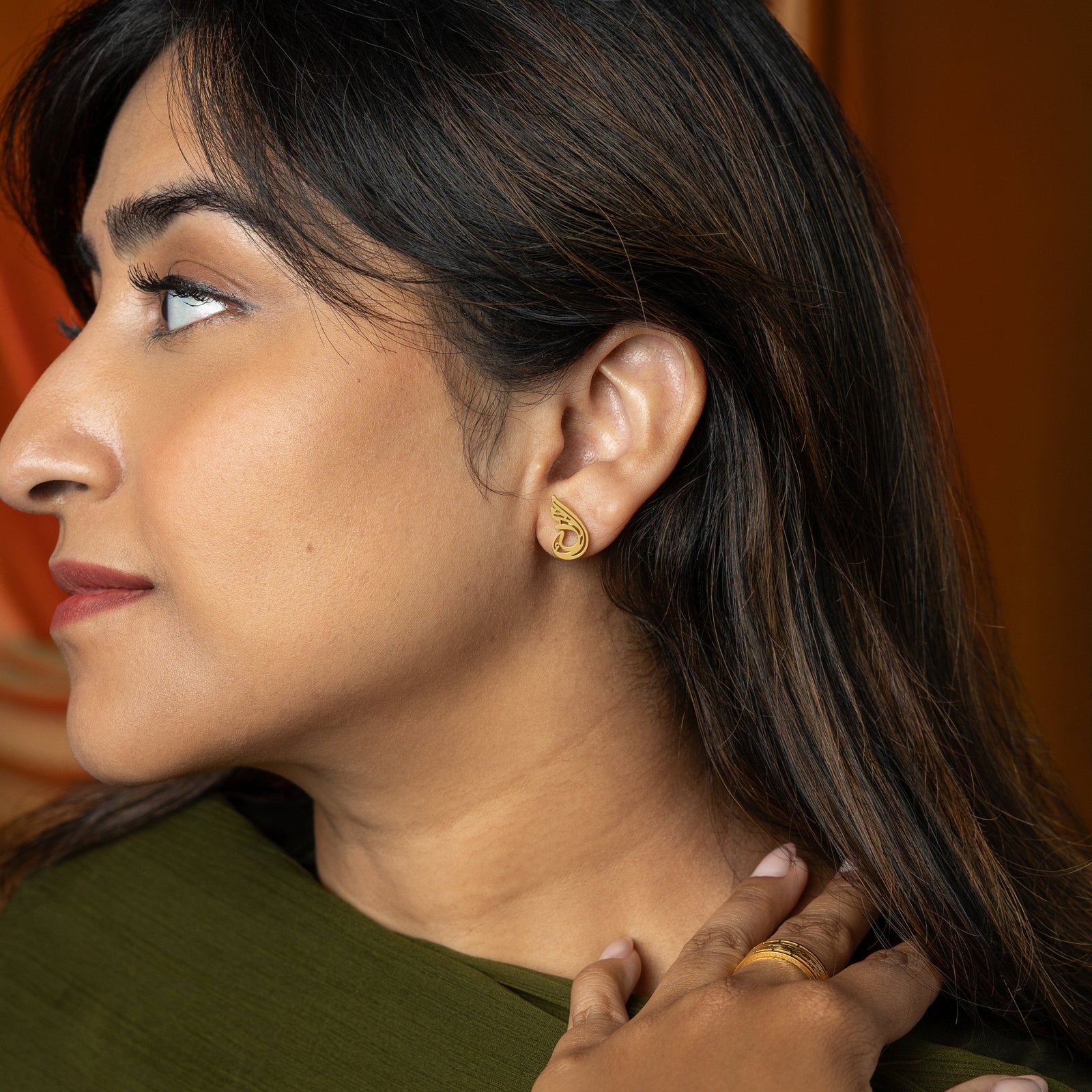 The image shows a person's ear adorned with a gold "Serenity Earring" from the Aadil x Nominal collection. The earring features a unique, abstract, teardrop-like design with swirling cut-out details. The metal has a warm gold color.