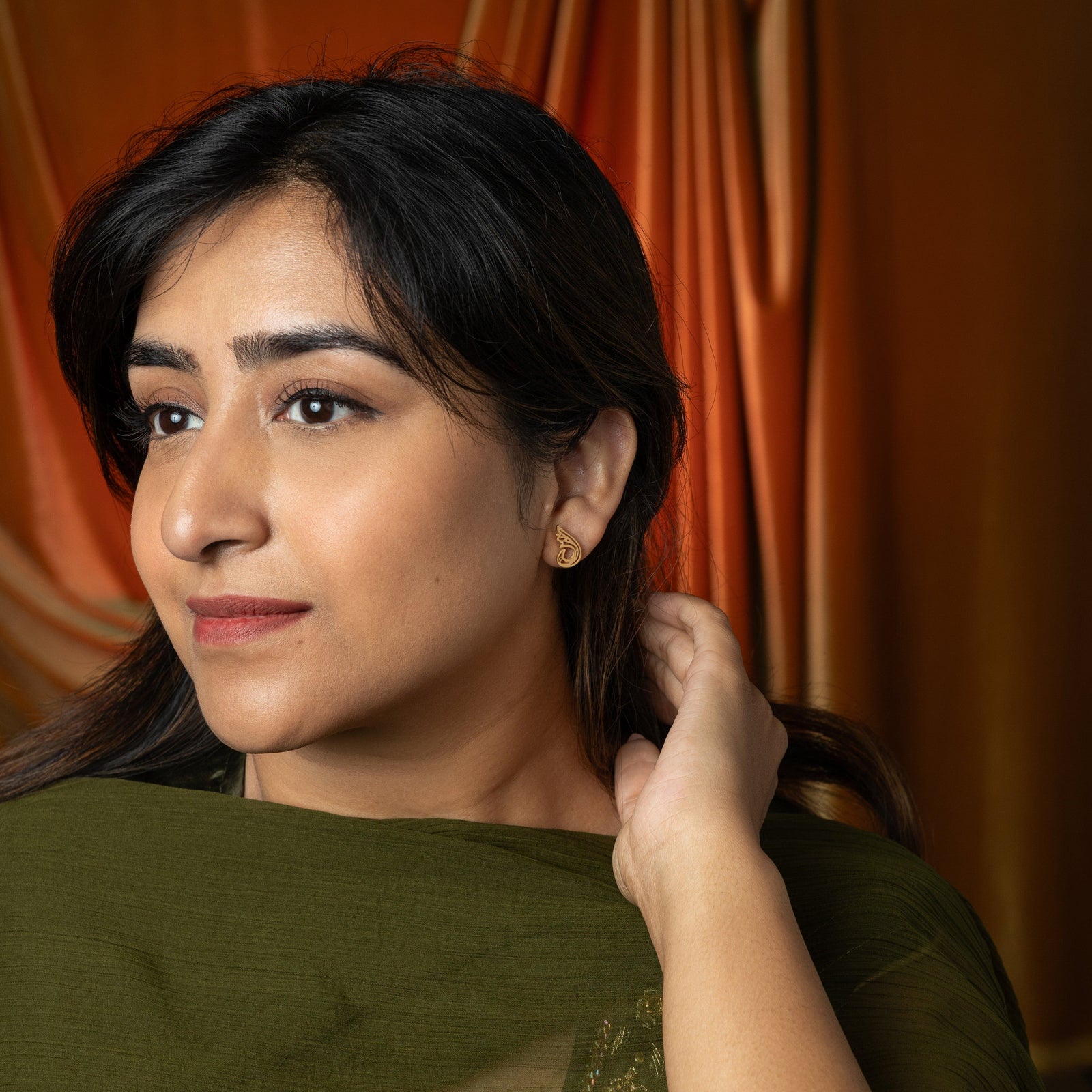 The image shows a person's ear adorned with a gold "Serenity Earring" from the Aadil x Nominal collection. The earring features a unique, abstract, teardrop-like design with swirling cut-out details. The metal has a warm gold color.
