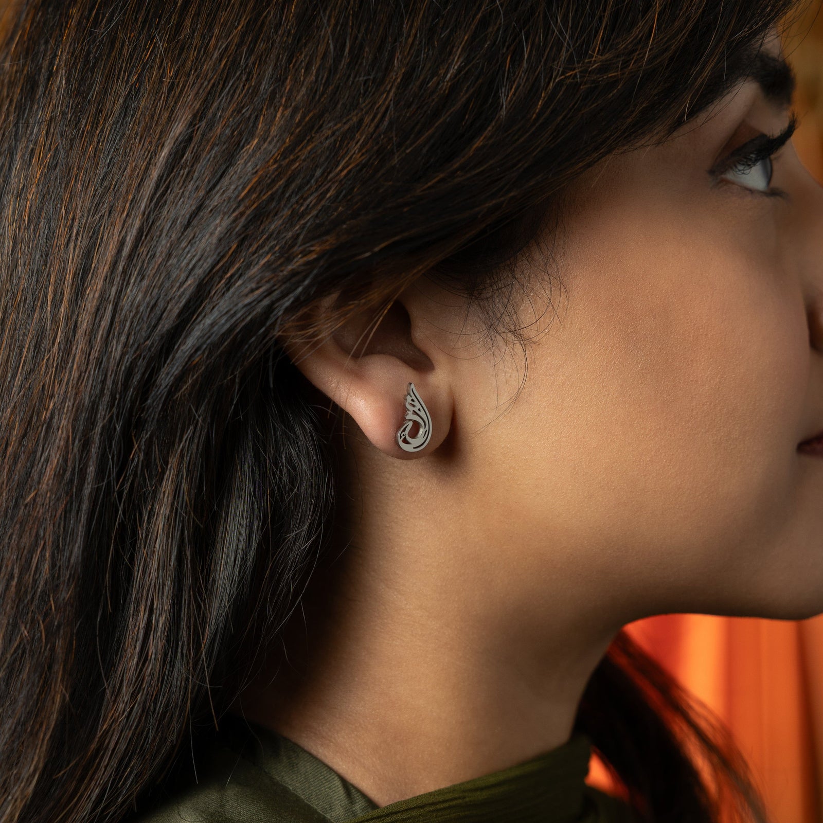 he image shows a person's ear adorned with a silver "Serenity Earring" from the Aadil x Nominal collection. The earring features a unique, abstract, teardrop-like design with swirling cut-out details. The metal has a light silver color. The person has dark hair and is wearing a dark green top.
