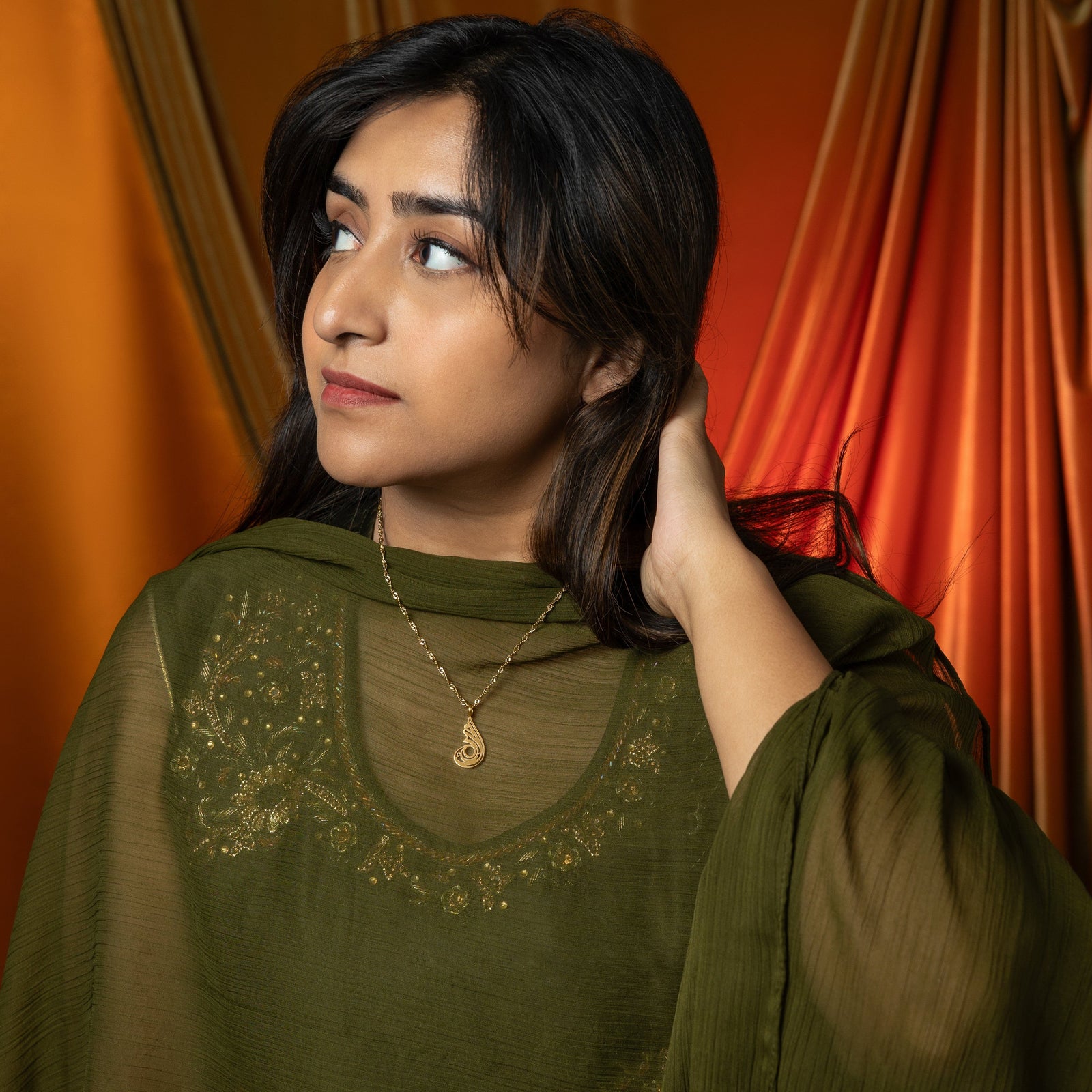 A delicate gold-plated chain necklace featuring a dainty, approximately 2 cm teardrop pendant with intricate, swirling cut-out details and a shiny finish. The necklace is worn by a person with dark hair and is shown against a blurred green garment. A hand with light-colored nail polish is gently holding the chain near the pendant.