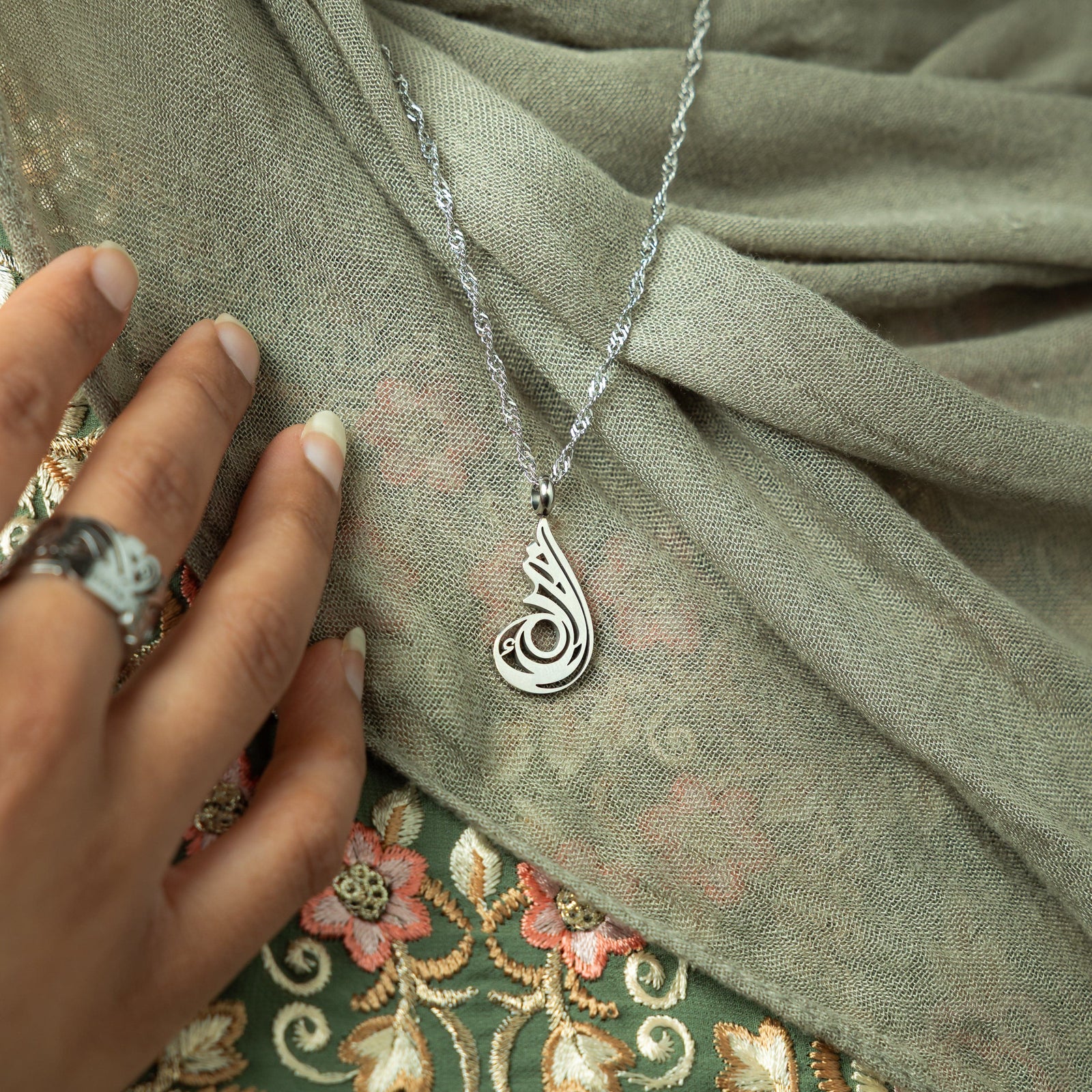 A delicate silver-plated chain necklace featuring a dainty, approximately 2 cm teardrop pendant with intricate, swirling cut-out details and a shiny finish. The necklace is worn against a blurred grey garment. A hand with a ring is gently holding the chain near the pendant.
