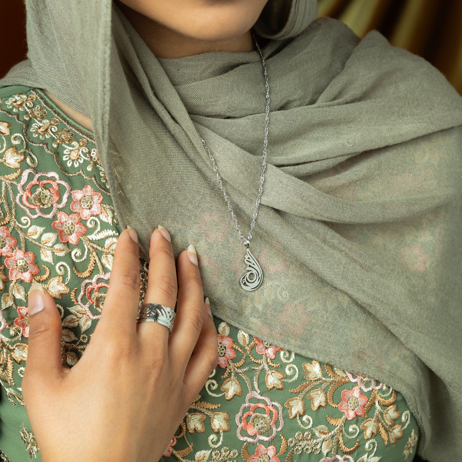 A delicate silver-plated chain necklace featuring a dainty, approximately 2 cm teardrop pendant with intricate, swirling cut-out details and a shiny finish. The necklace is worn against a blurred grey garment. A hand with a ring is gently holding the chain near the pendant.