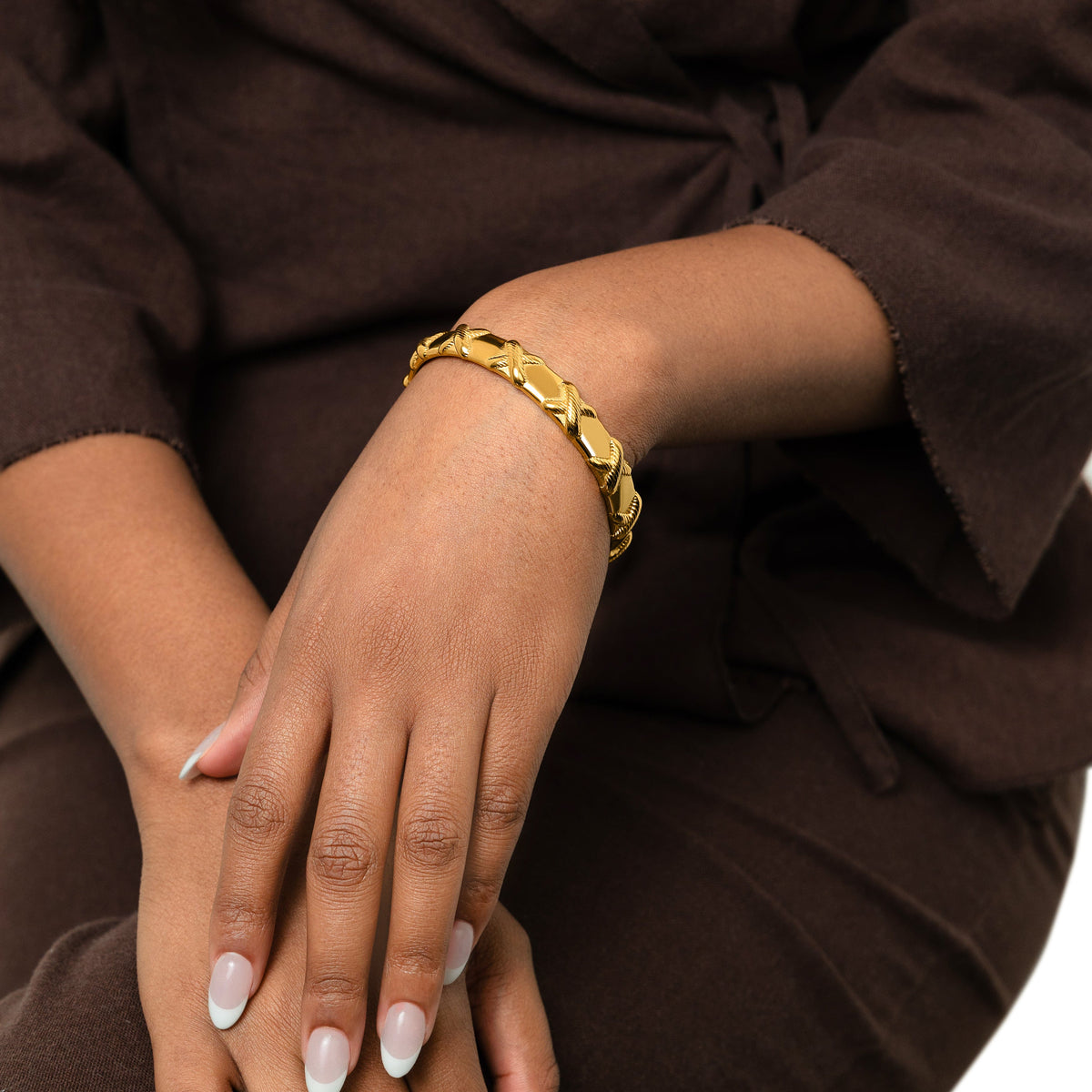Photo of the model's hands on their lap. The model is wearing the gold "Leave Your Mark | X" Bangle on their left wrist.