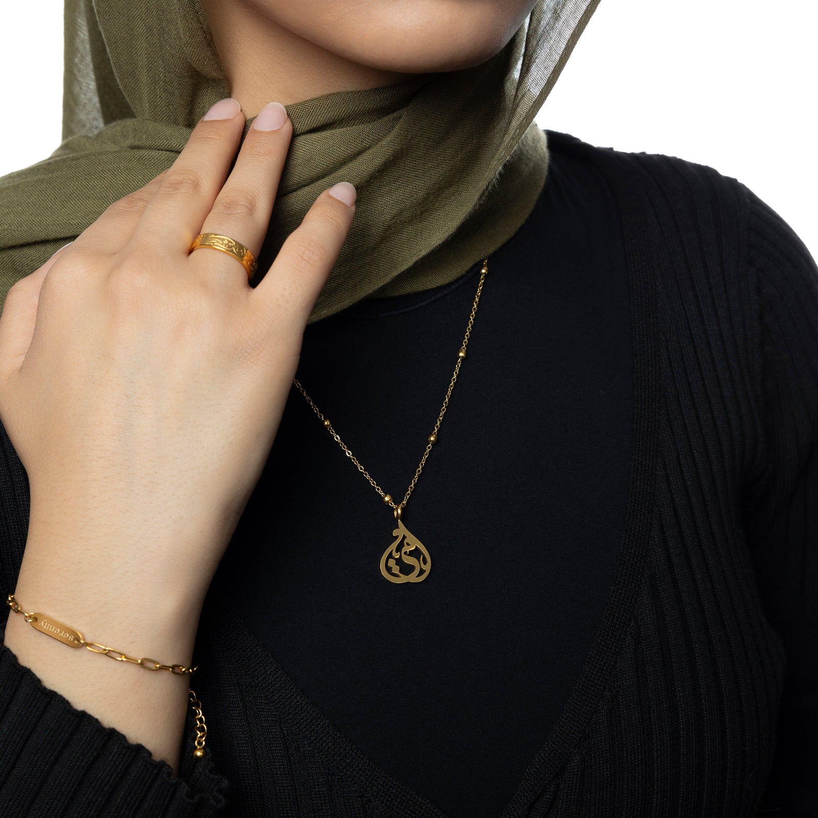 Woman wearing a gold Sister Calligraphy Necklace layered over a black top and styled with a matching gold ring and bracelet, olive green hijab, and hand resting near neckline.