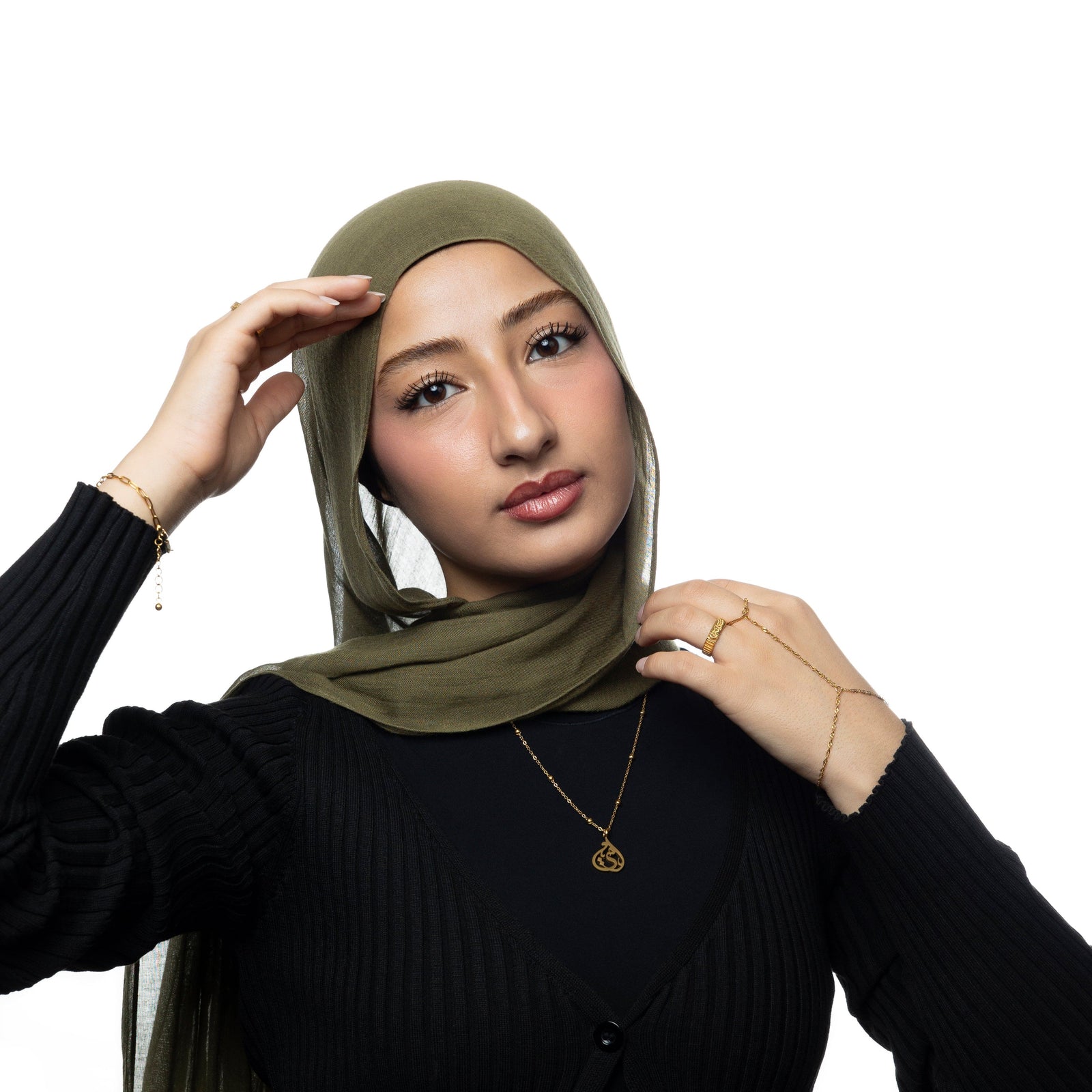 Woman wearing full gold jewelry set including the Sister Calligraphy Necklace, styled with an olive green hijab and black ribbed cardigan. Necklace rests at center of chest.