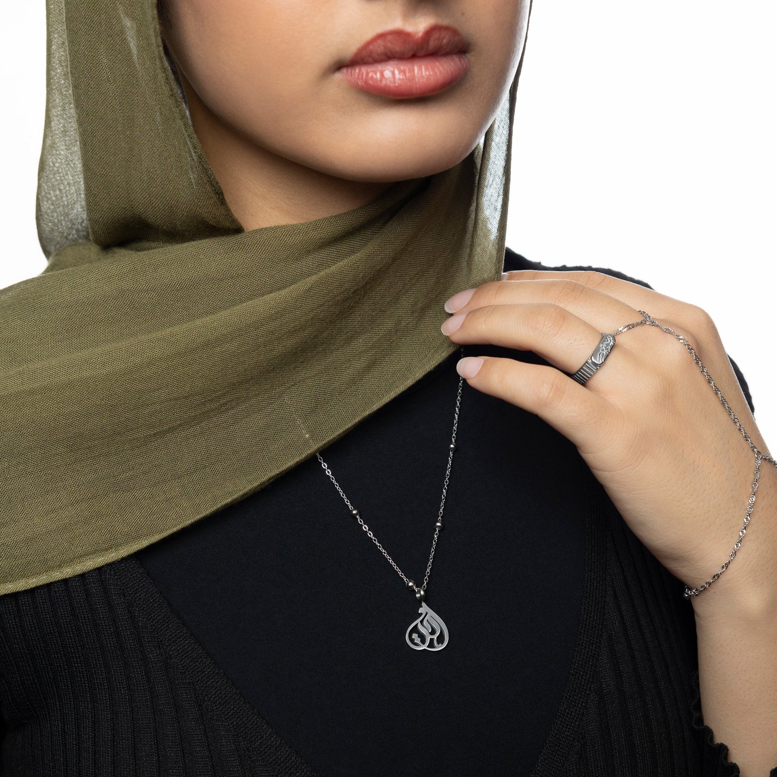 Woman wearing silver Sister Calligraphy Necklace over a black top, paired with a silver ring and silver hand chain, styled with olive green hijab.