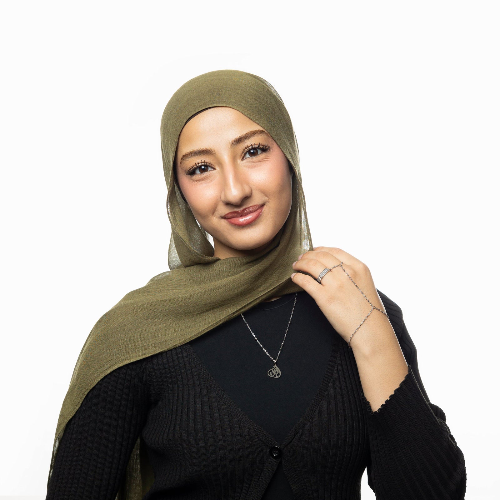 woman wearing silver Sister Calligraphy Necklace and coordinating silver jewelry, dressed in black with olive green hijab. Necklace pendant with Arabic word “Sister” rests on chest.