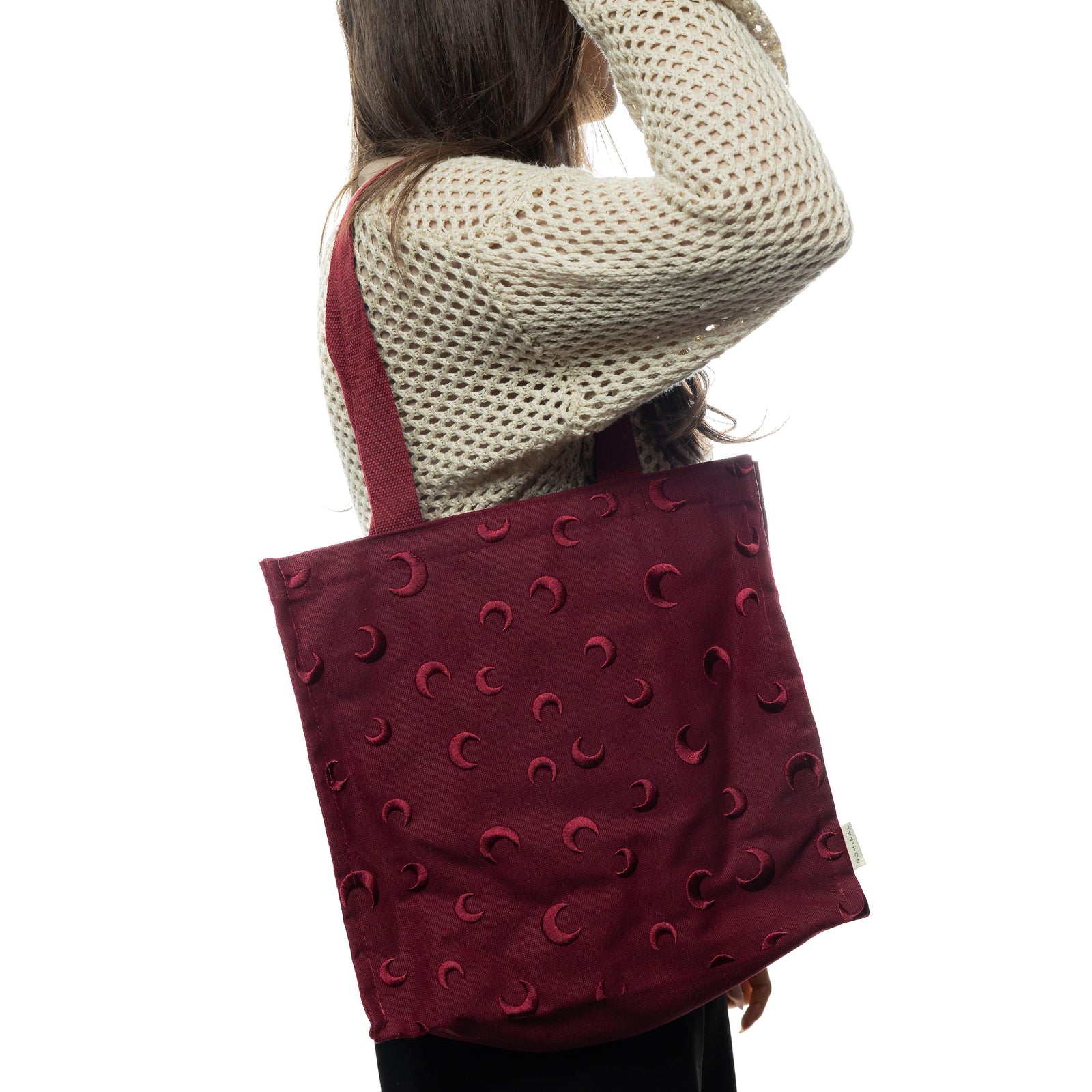 Woman wearing a cream knit sweater carrying the Skyfall Tote Bag on her shoulder. The bag features tonal maroon crescent moon embroidery throughout.