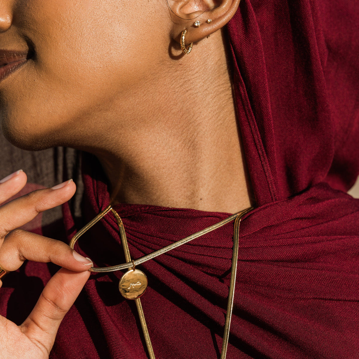 Close up of the gold Sabr / Patience Charm as worn by the model. The model lifts the chain to show the gold pendant.