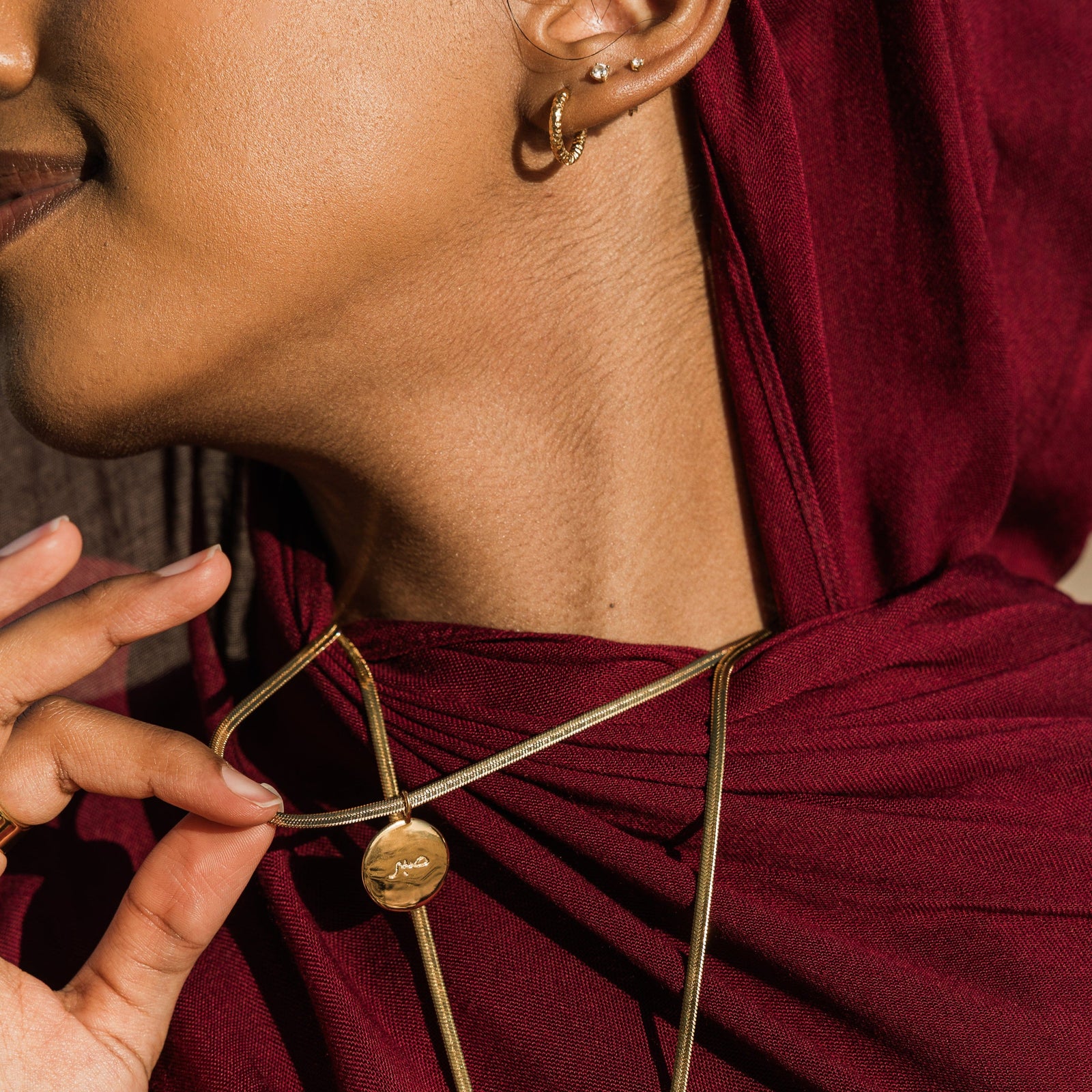 Close up of the gold Sabr / Patience Charm as worn by the model. The model lifts the chain to show the gold pendant.
