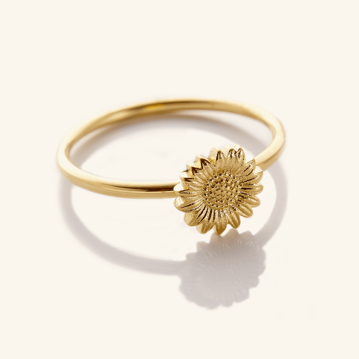 Close-up of a thin gold ring featuring a detailed sunflower design on top, photographed against a light background.