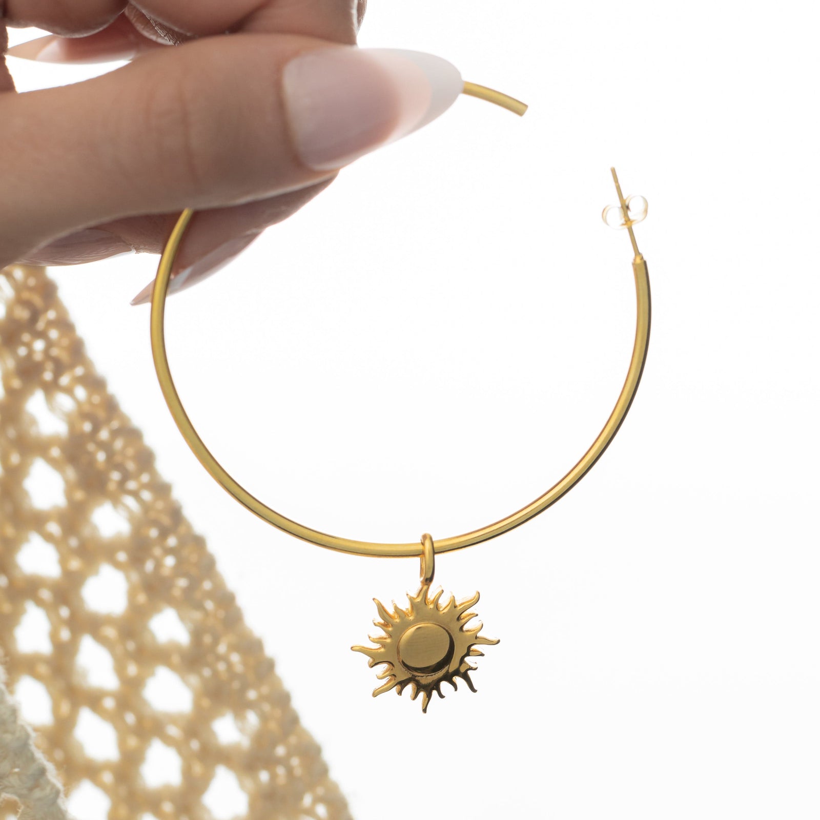 Gold hoop earring with a dangling sunshine charm featuring spiked rays and a round center, held between fingers in front of a white backdrop with beige crochet fabric in the background.