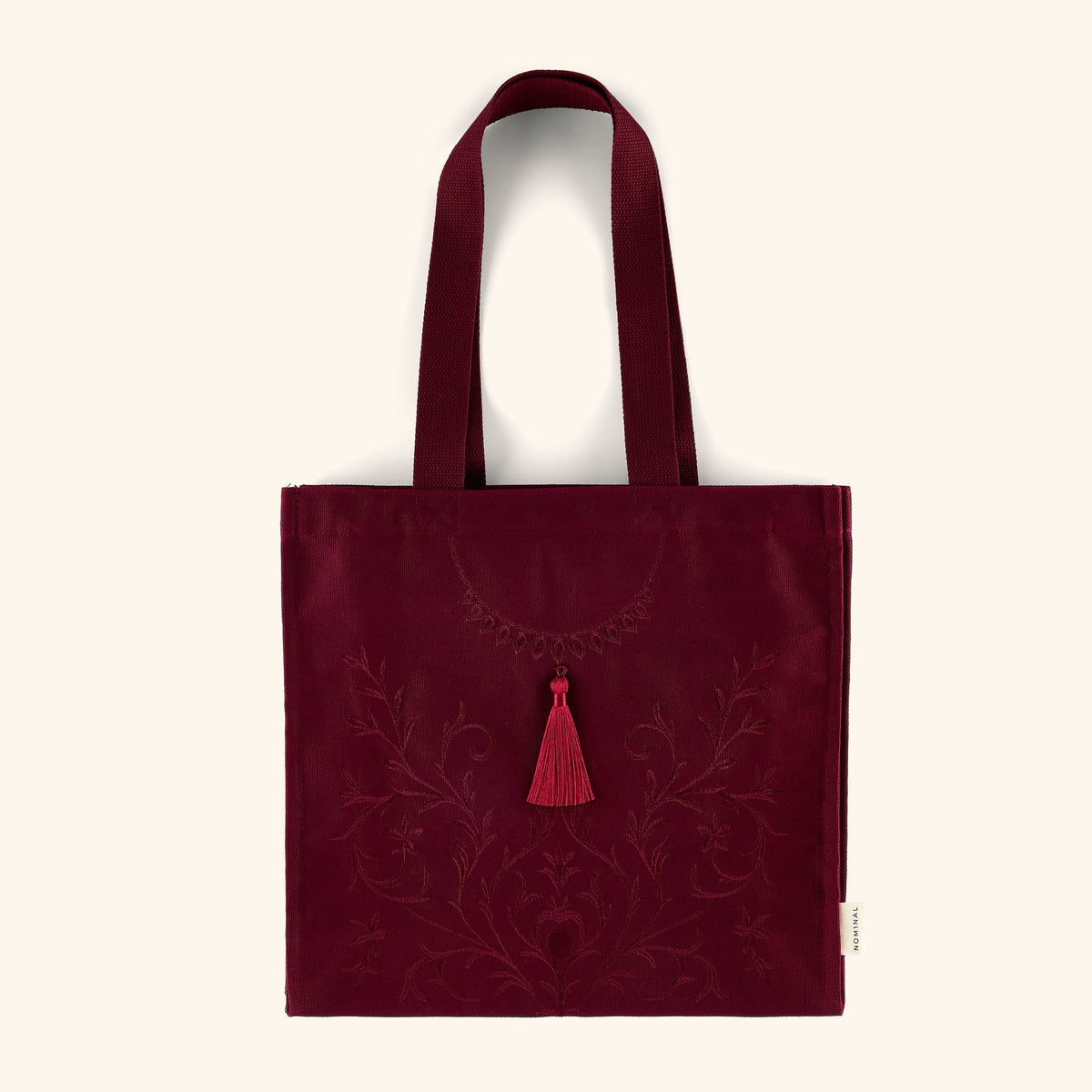 A maroon textured cotton canvas tote with a matching embossed embroidery that features florals swirling around the canvas finished with a hanging tassel.