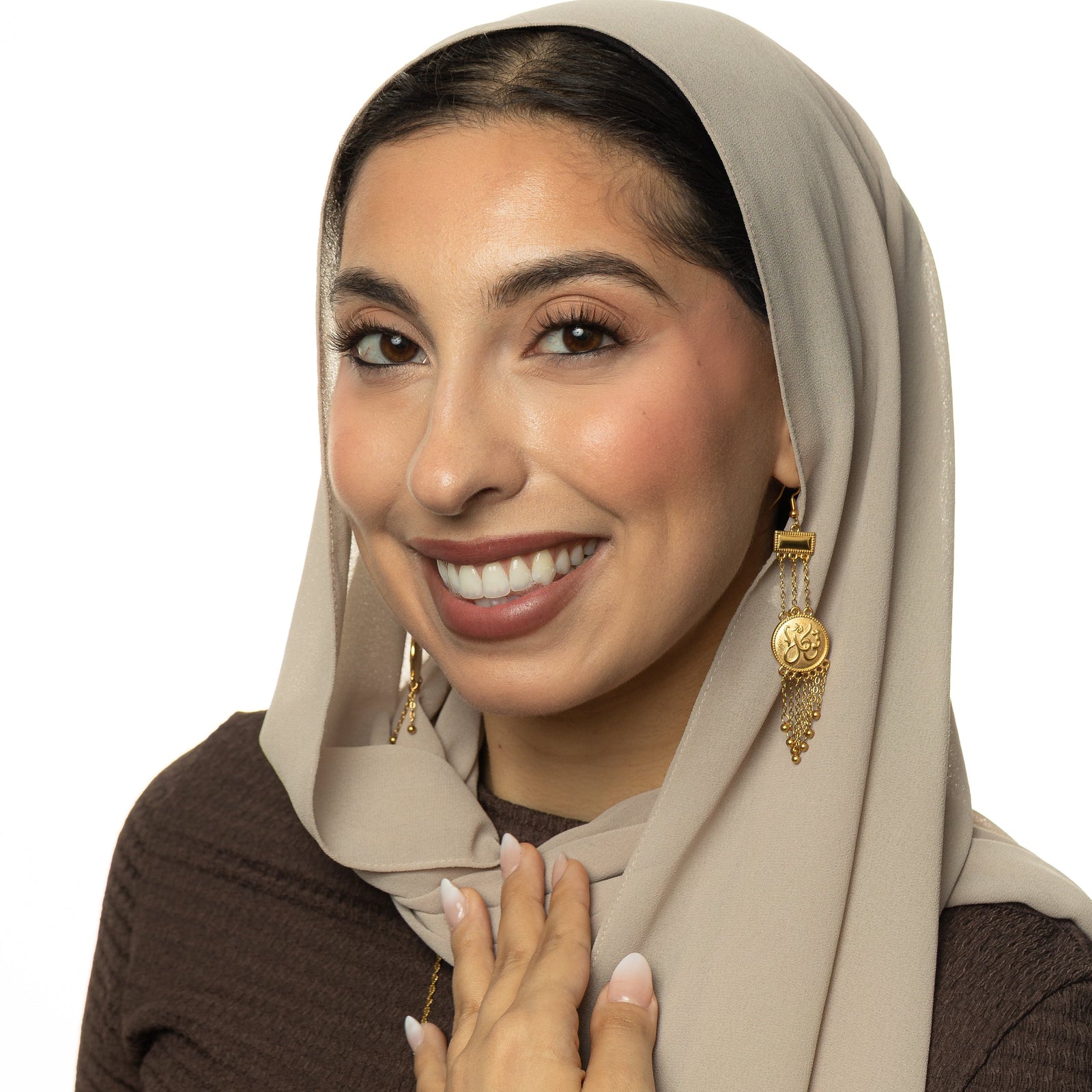 Photo of a model wearing a light beige headscarf draped neatly around her head and shoulders, paired with a dark brown top. Model is wearing the gold Tawakkul Trust Raining Earrings.