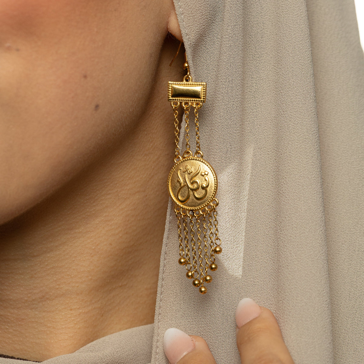 Gold Tawakkul | Trust Raining Earrings shown worn, featuring circular coin-style pendants engraved in Arabic calligraphy with multiple dangling gold chains and small bead drops.