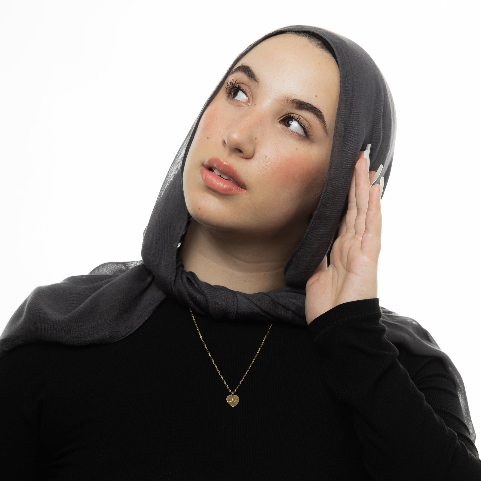 Photo of the model looking upward and touching their hijab with their left hand. the model is wearing the gold Trust | توكل Charm Necklace.