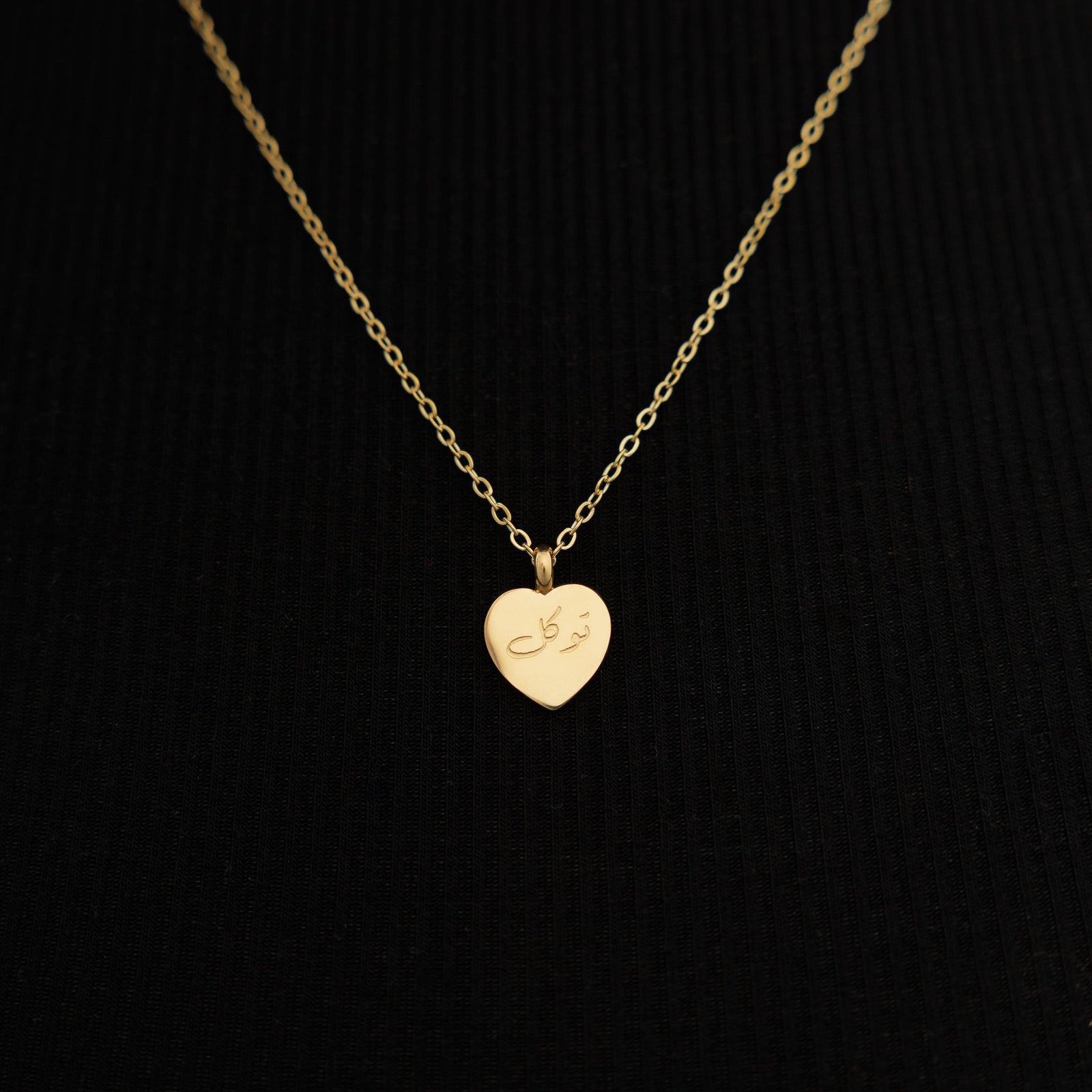 Close up of the gold Trust | توكل Charm Necklace against the black fabric of the model's clothing.