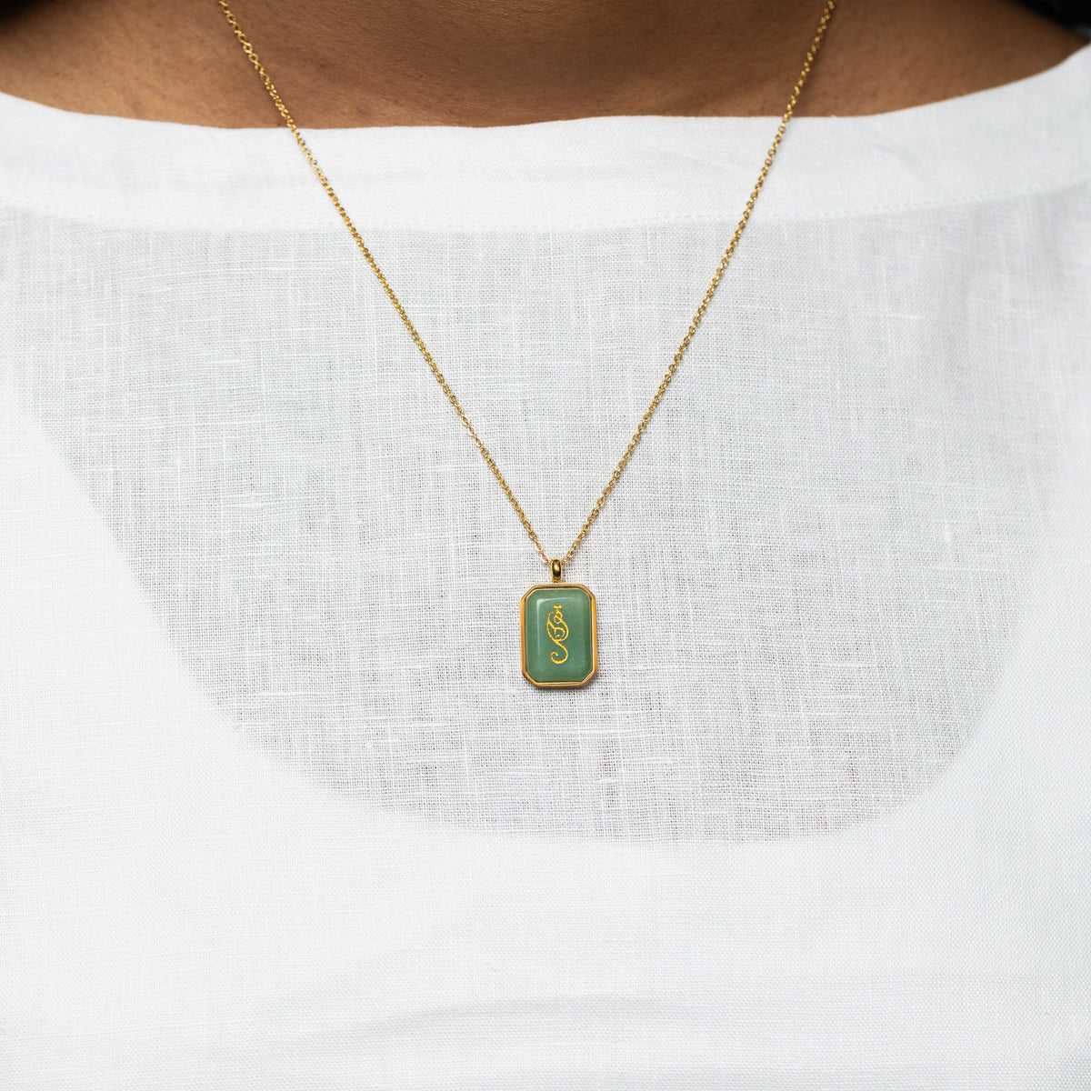 Close-up of a woman wearing a gold chain necklace with a rectangular green aventurine pendant engraved with "Tawakkul" in Arabic calligraphy, styled with a white textured blouse.