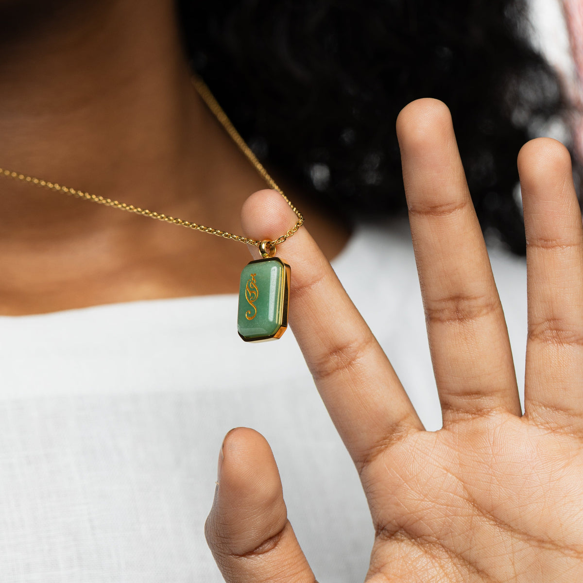 Woman holding a gold chain necklace with a rectangular green aventurine pendant engraved with "Tawakkul" in Arabic calligraphy, styled against a white blouse.