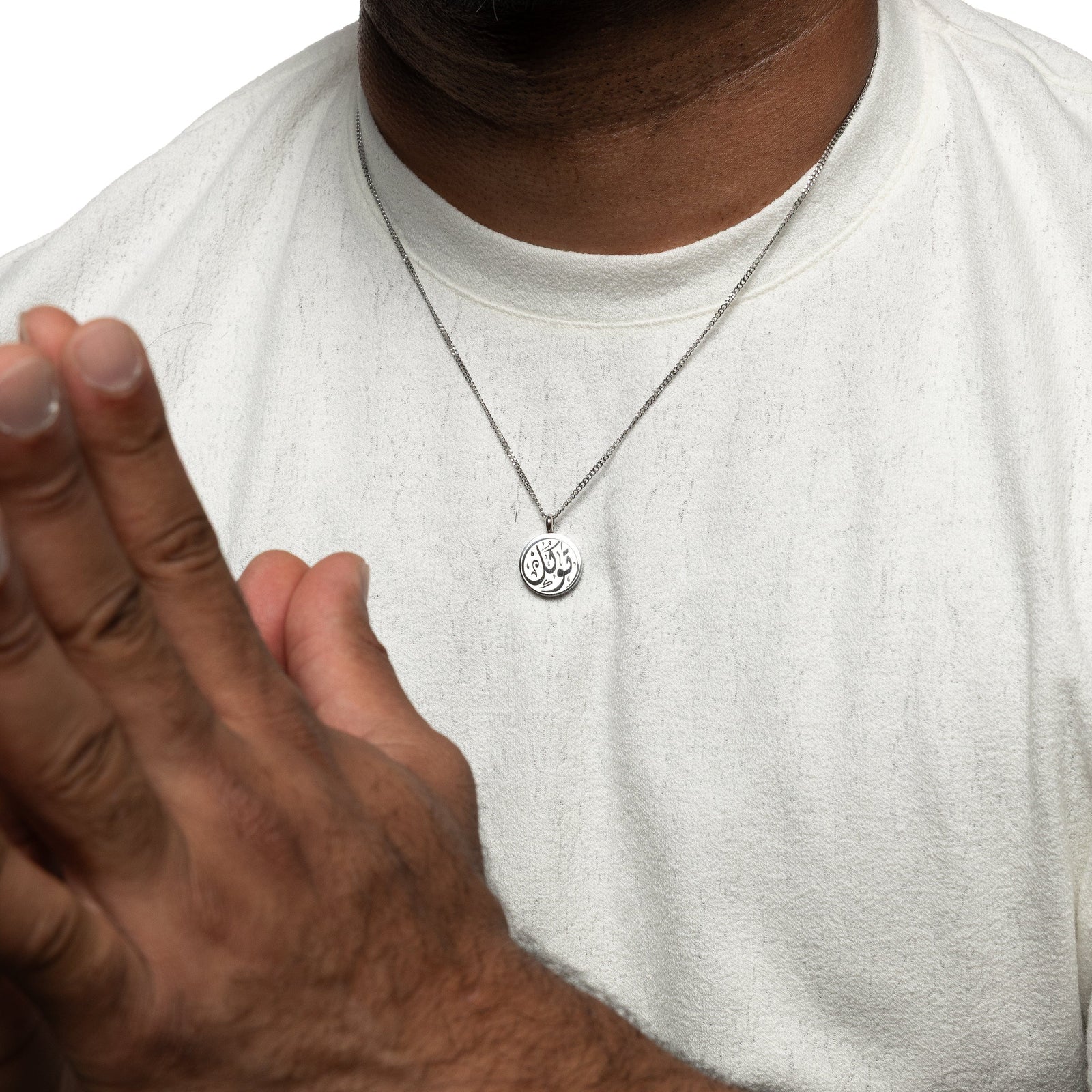Silver necklace with circular pendant engraved with 'Tawakkul' calligraphy, worn over a white shirt.