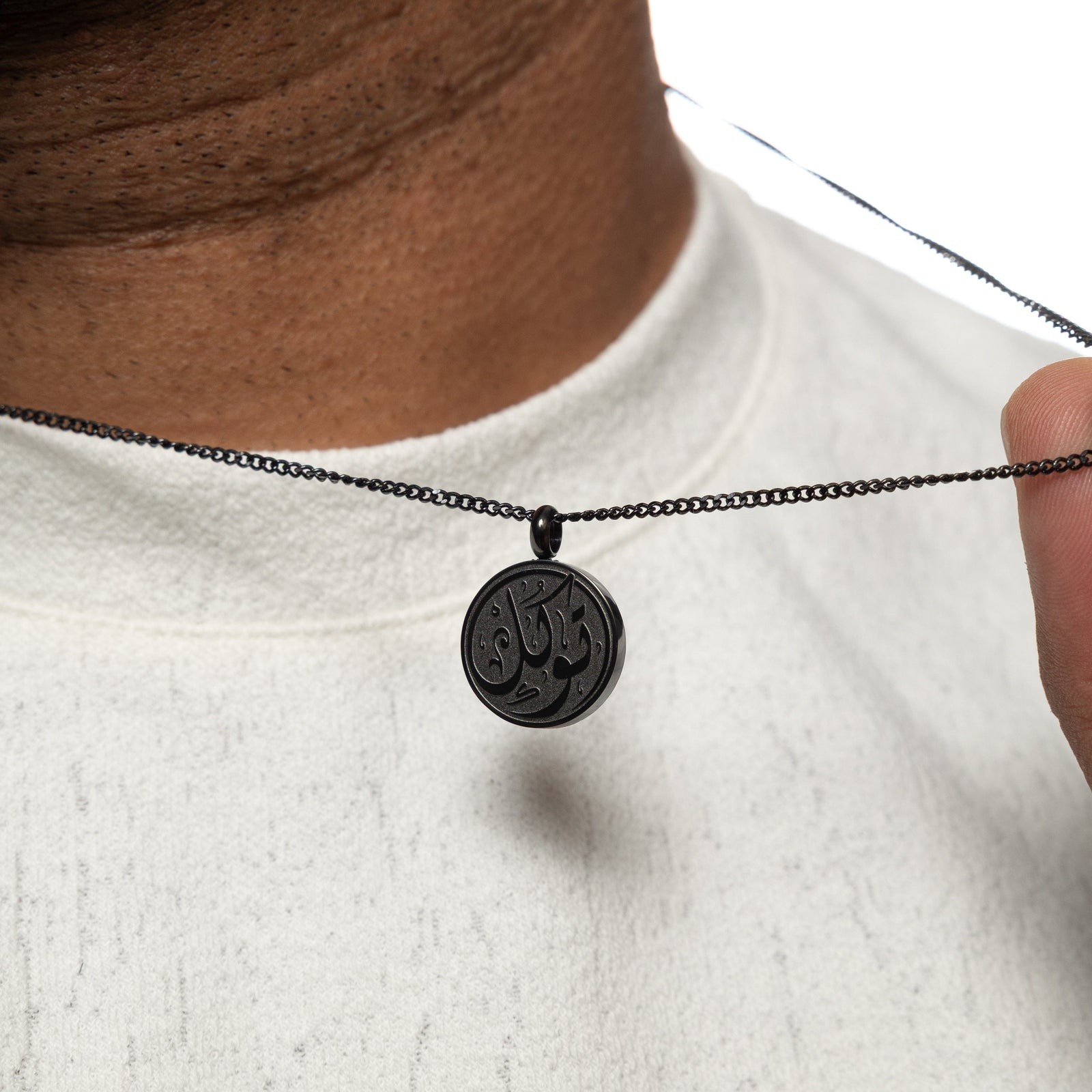 Close-up of a man holding up a black necklace with a circular pendant engraved with 'Tawakkul' calligraphy, worn over a white shirt.