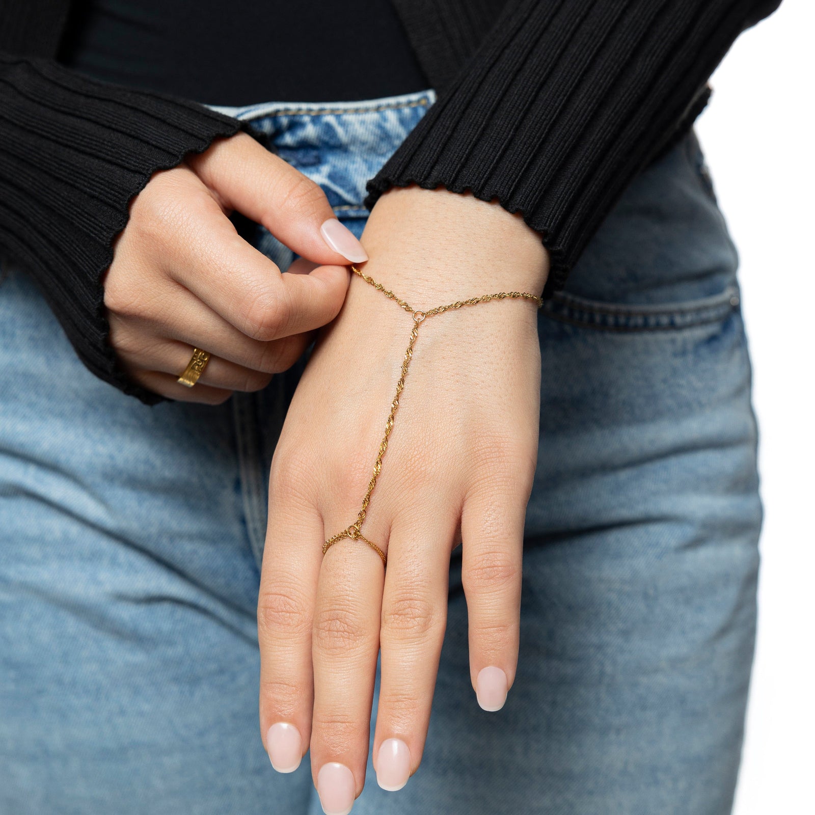 Woman wearing a gold twisted hand chain, photographed against light blue jeans, showing chain looping from wrist to middle finger.